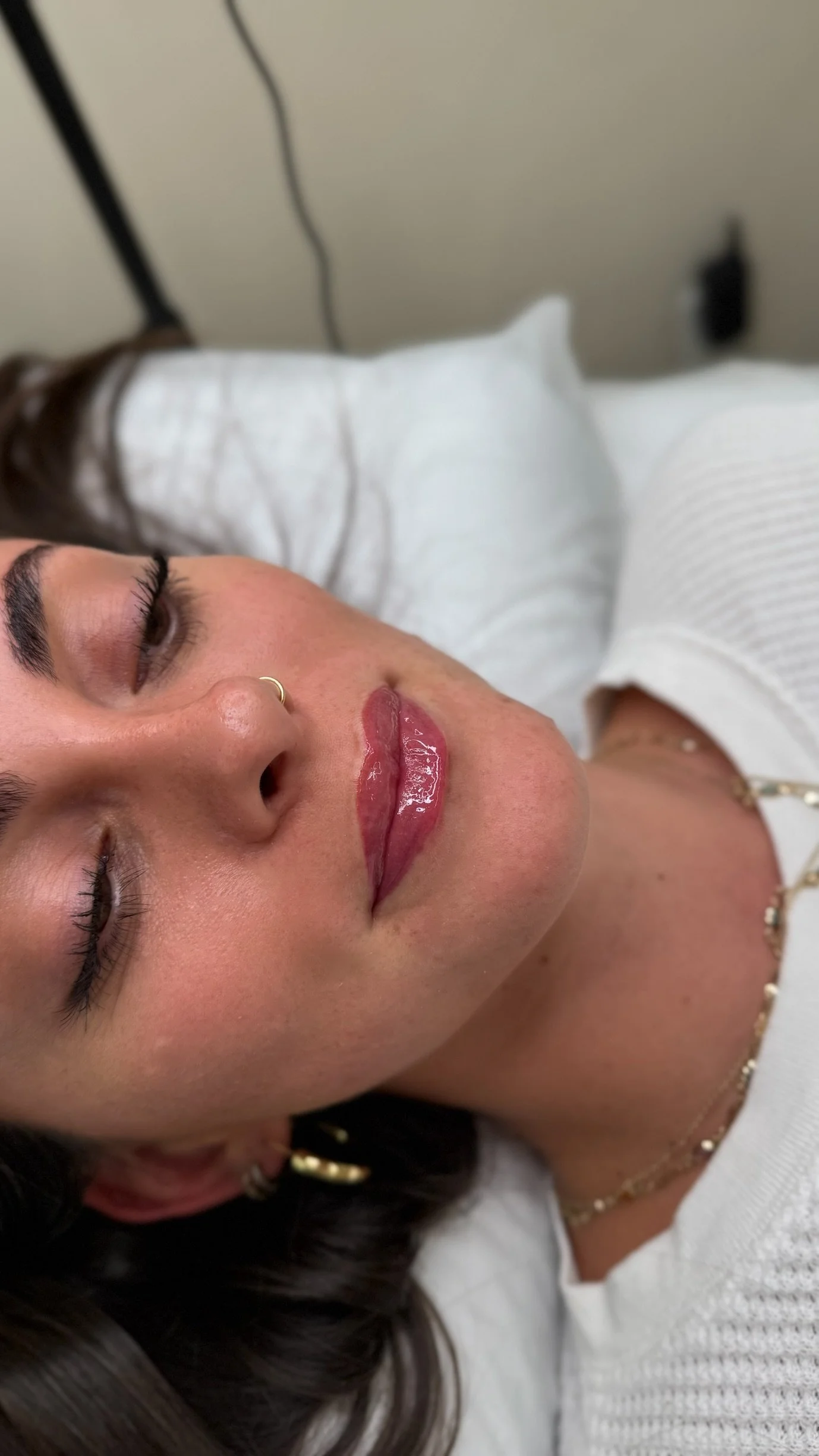 Close-up of a woman with closed eyes lying on a bed, showing her face with shiny, pink lipstick, a small nose ring, and layered necklaces, wearing a white top.