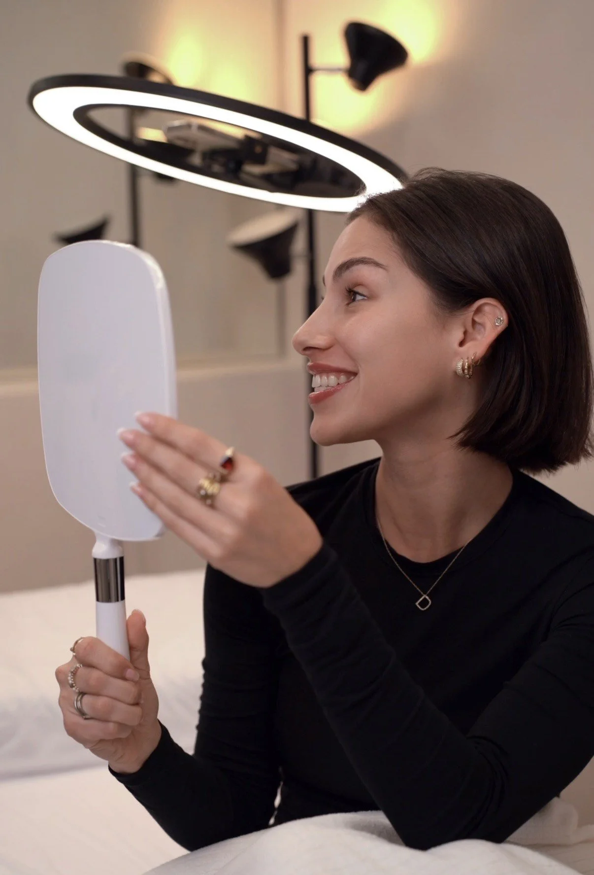 A woman with short dark hair and earrings looks at herself in a mirror, smiling, while holding a handheld mirror in her hand, with a ring on her finger.