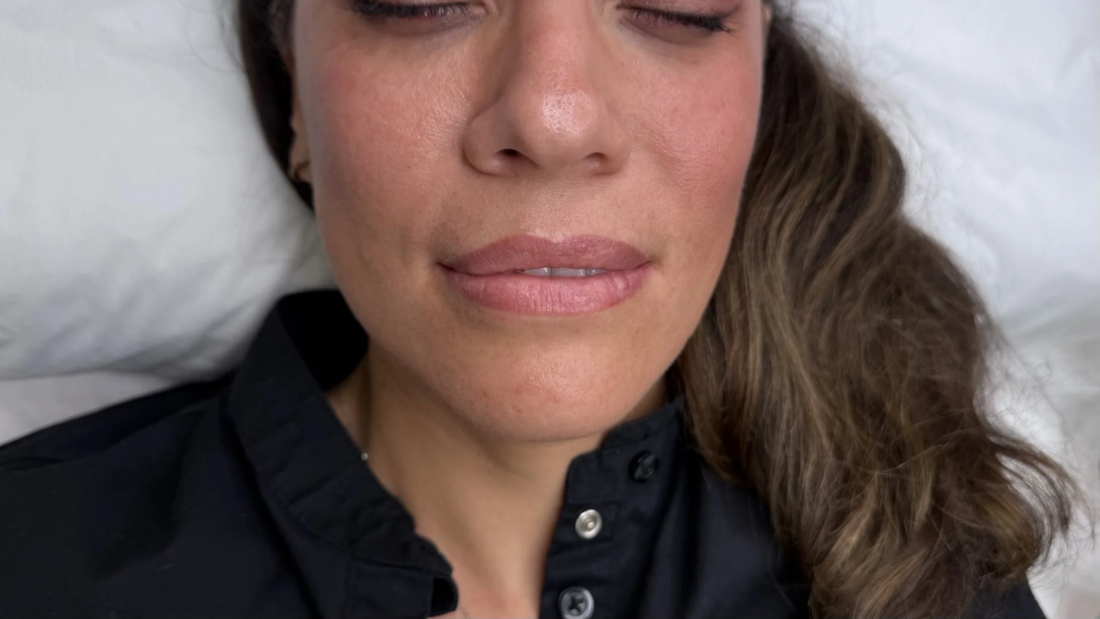 Close-up of a woman with closed eyes and light makeup, lying on a white pillow, wearing a black shirt.