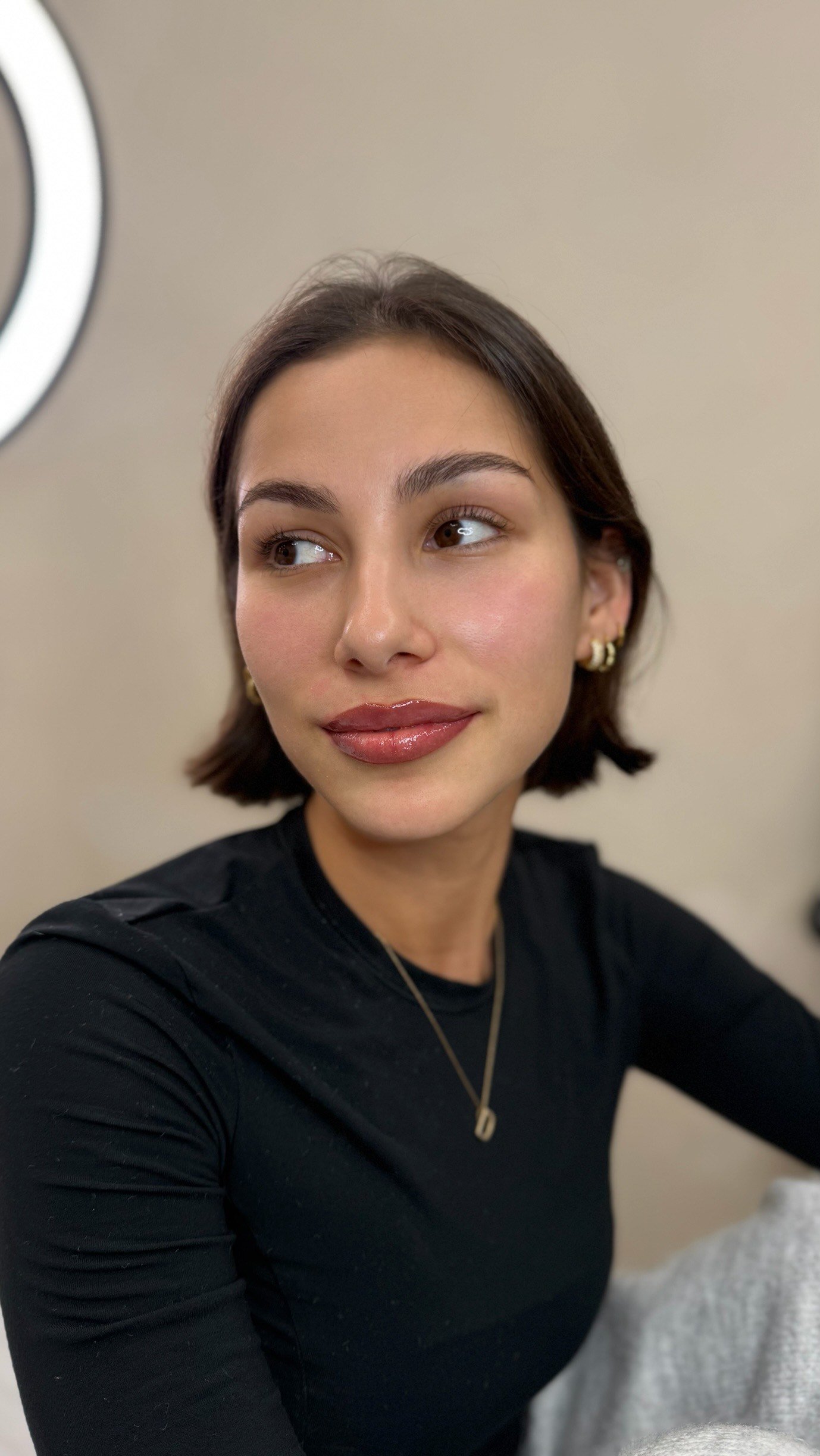 Close-up of a young woman with short brown hair, wearing a black top, gold jewelry, and lipstick, looking slightly to her right, in front of a beige background with part of a ring light visible.