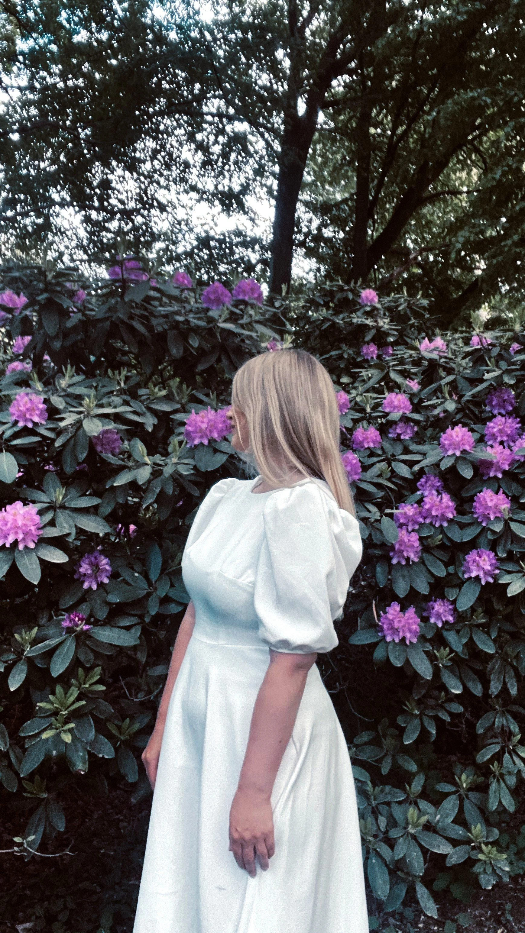Eine Frau mit blondem Haar trägt ein weißes Kleid und steht vor einem Busch mit pinken Blüten. Im Hintergrund sind große Bäume und Himmel sichtbar.