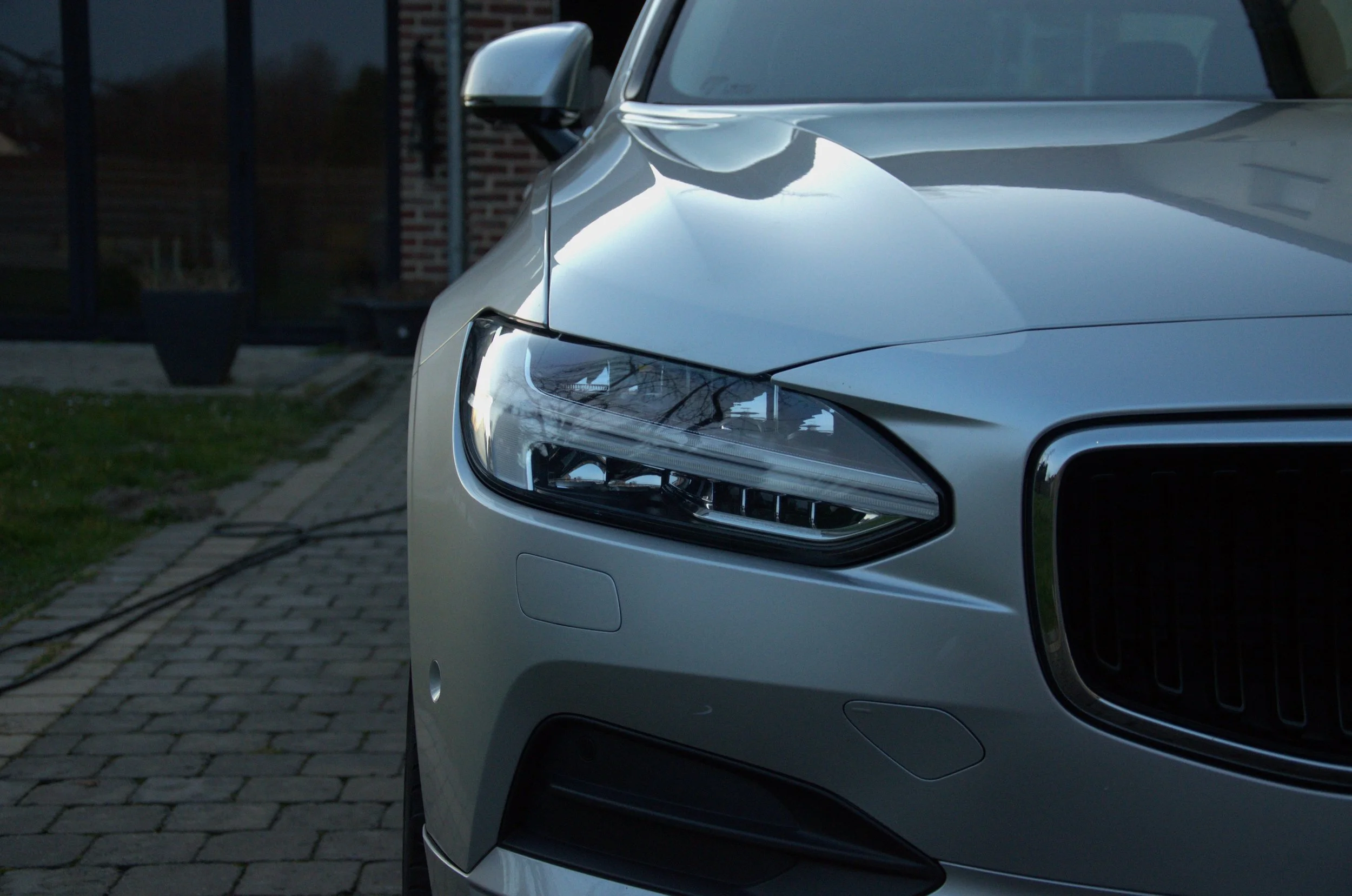 Dichtbij portier van een zilveren auto met een futuristisch ontwerp, met duidelijke koplampen en spiegel, geparkeerd op een straat met stenen bestrating, nabij een gebouw met een bakstenen muur.