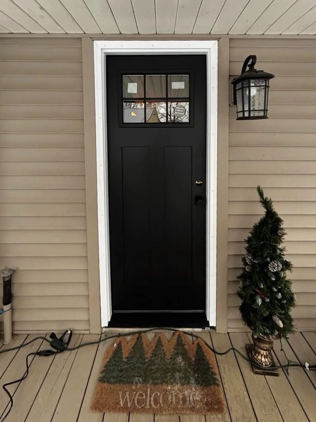 Front porch with black door, white framing, small holiday tree in a decorative planter, welcome mat with trees, and a wall-mounted outdoor lantern light