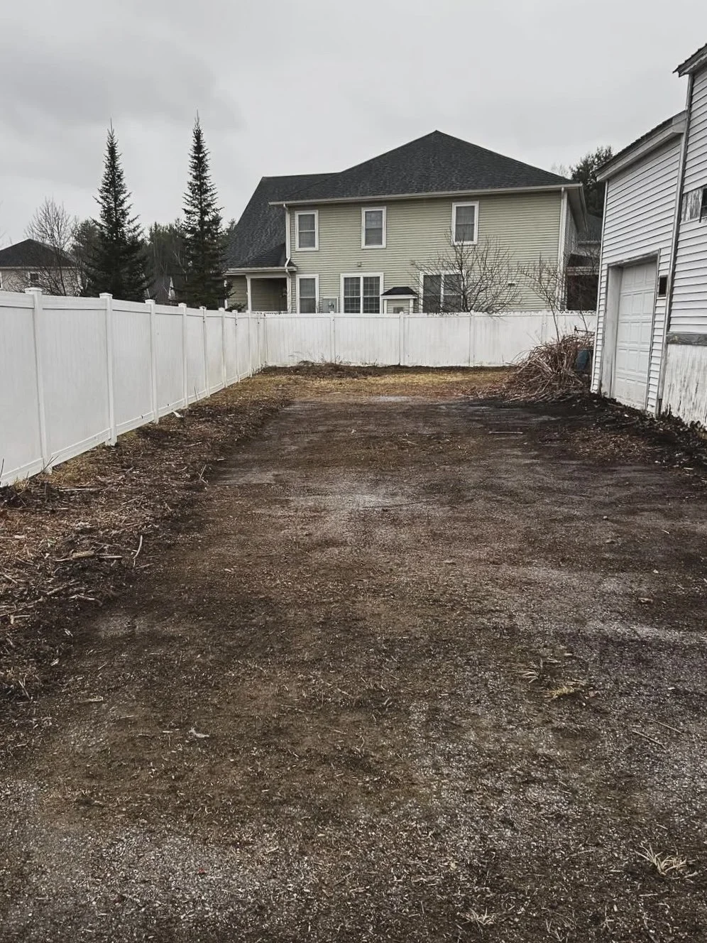 A backyard with a dirt and gravel surface, enclosed by a white fence, with a two-story house and trees in the background on a cloudy day.