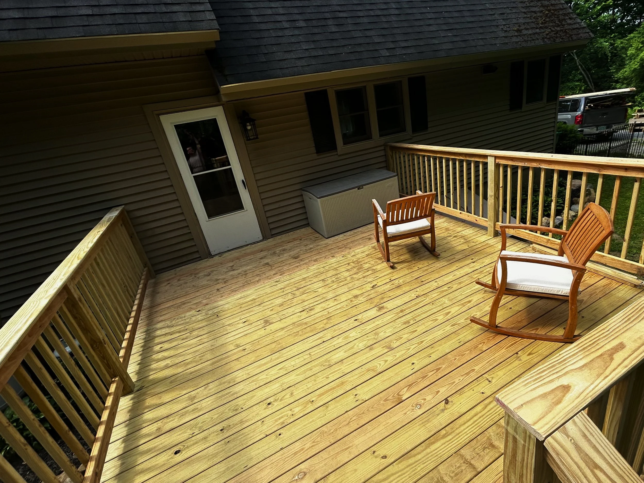 Image of a wooden deck attached to the back of a house, with two rocking chairs with white cushions, a storage box, and a door leading inside. The house has beige siding and black shutters, with trees and a car visible in the background.