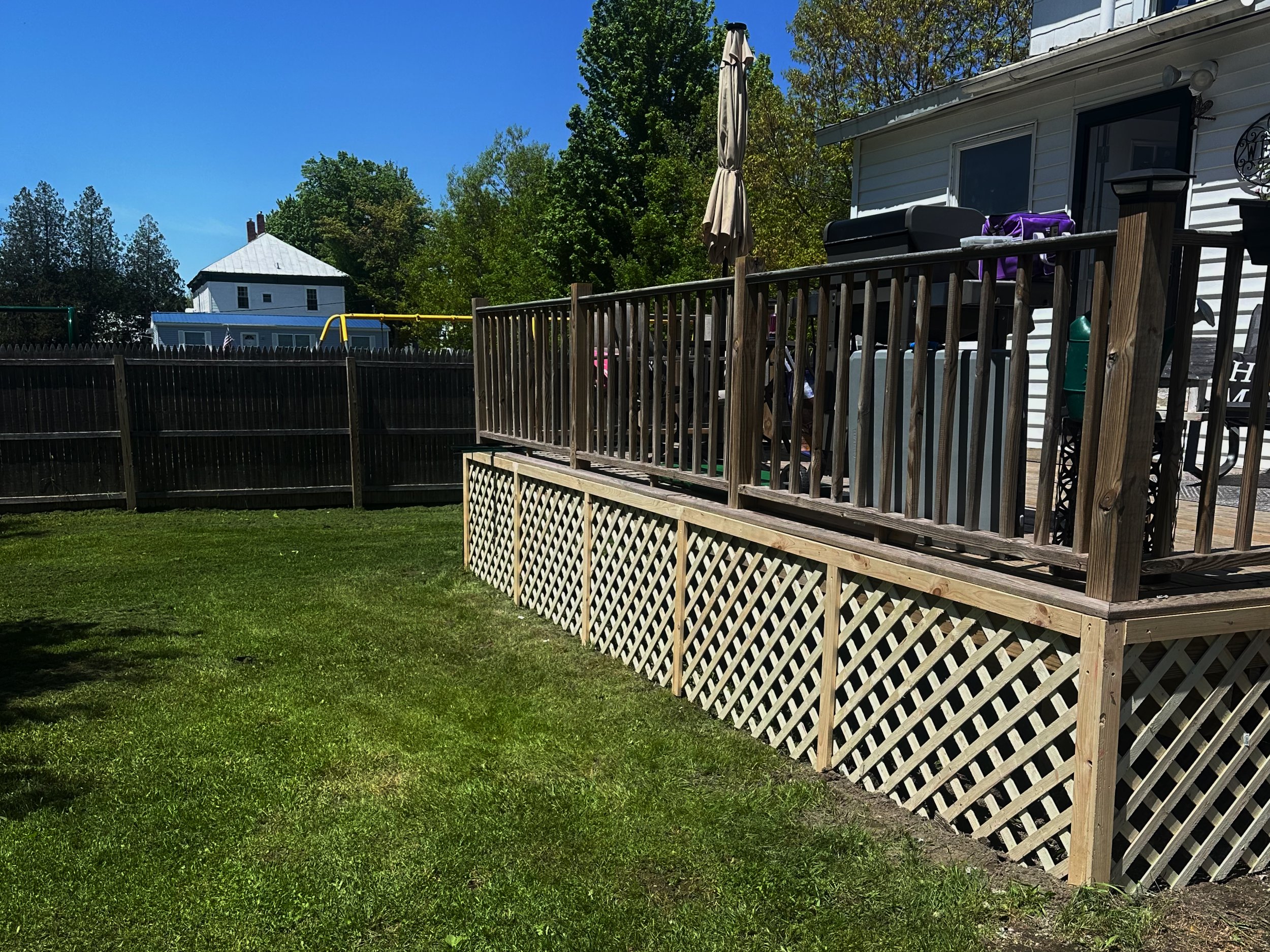 A backyard with a green lawn, wooden deck with railing and lattice skirting, and a white house. There is a closed umbrella and some outdoor furniture on the deck. A black wooden fence surrounds the yard, and there are trees and houses visible in the 