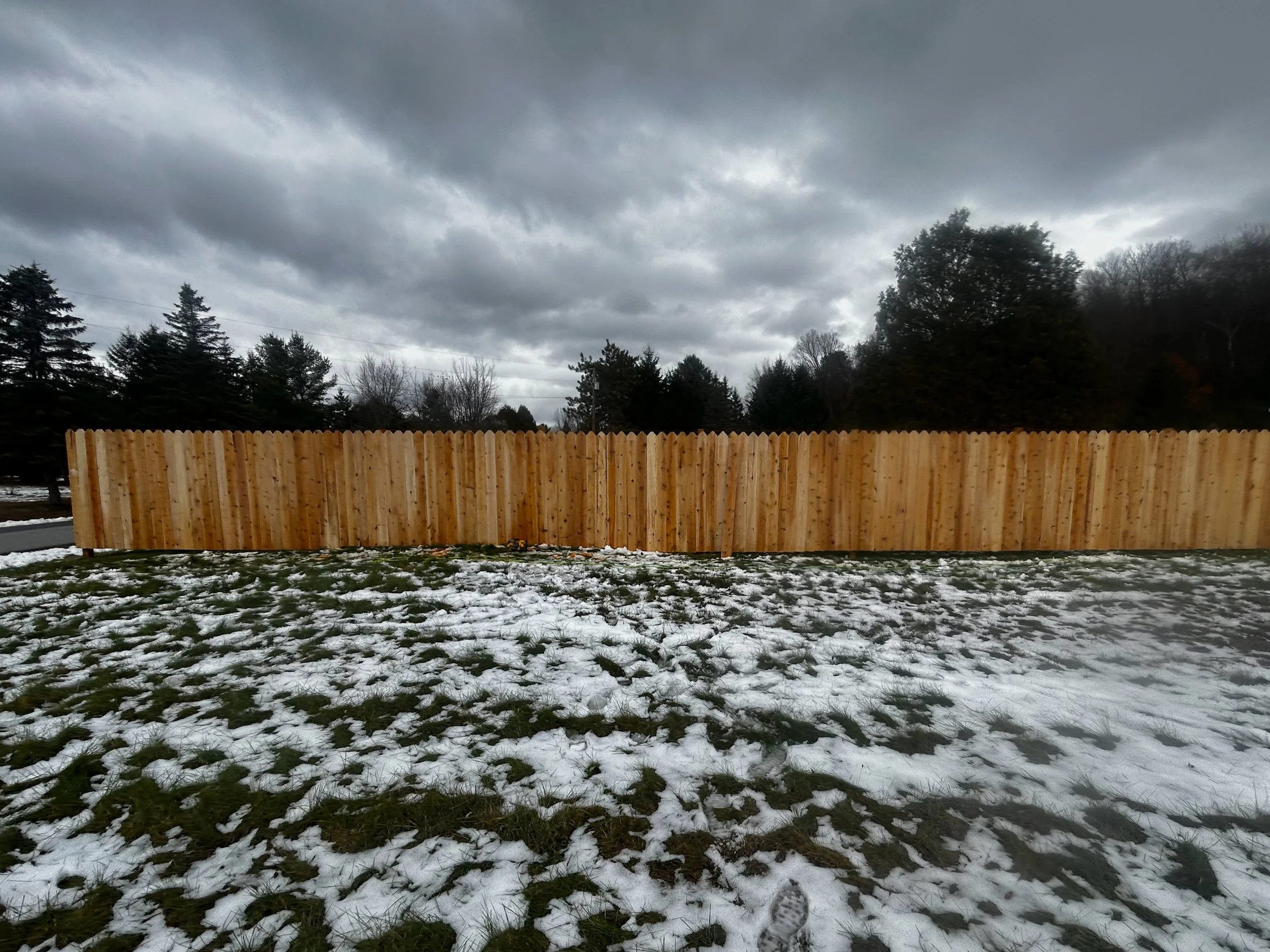 A newly constructed wooden fence on a patchy snow-covered yard with overcast sky and trees in the background.