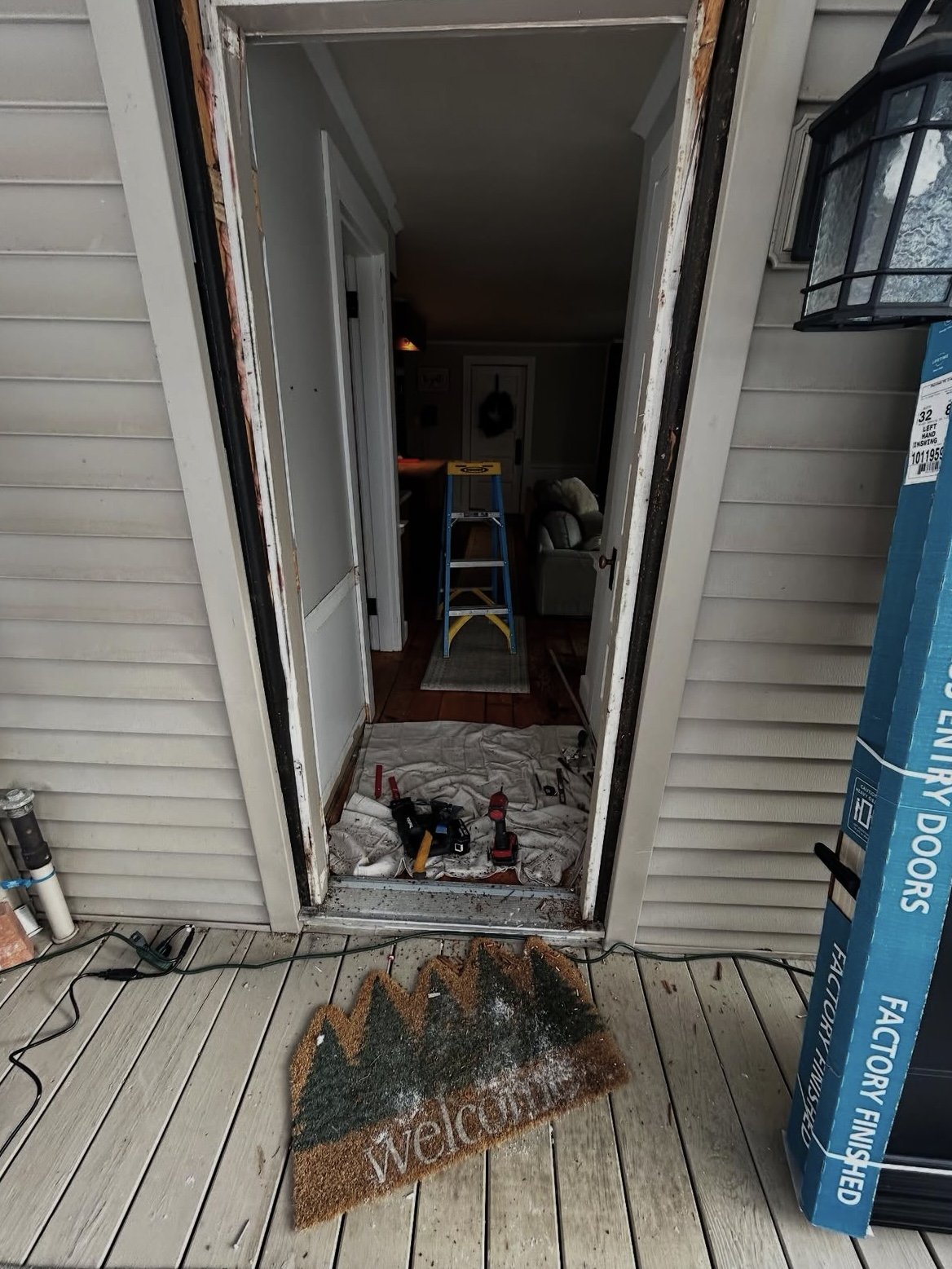 The photo shows a front door under renovation, with the door frame removed. Outside, there is a welcome mat on the porch, next to construction tools and a box of new storm doors. Inside, a blue ladder and a sofa are visible in a room with wooden floo
