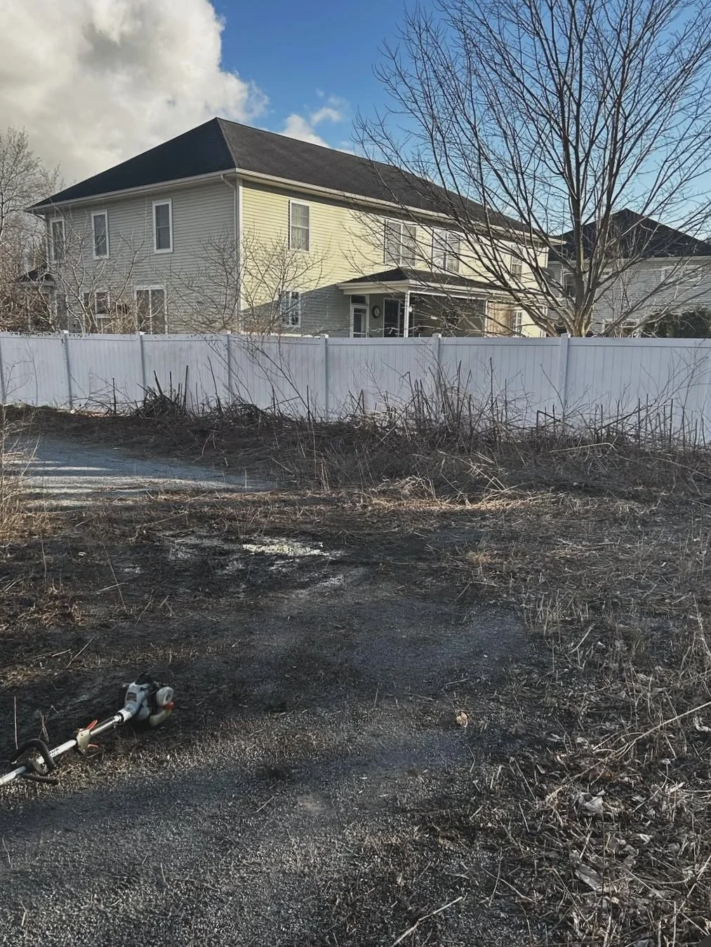 Backyard with bare trees, a white fence, and a burned area in the foreground. A power tool is on the ground.