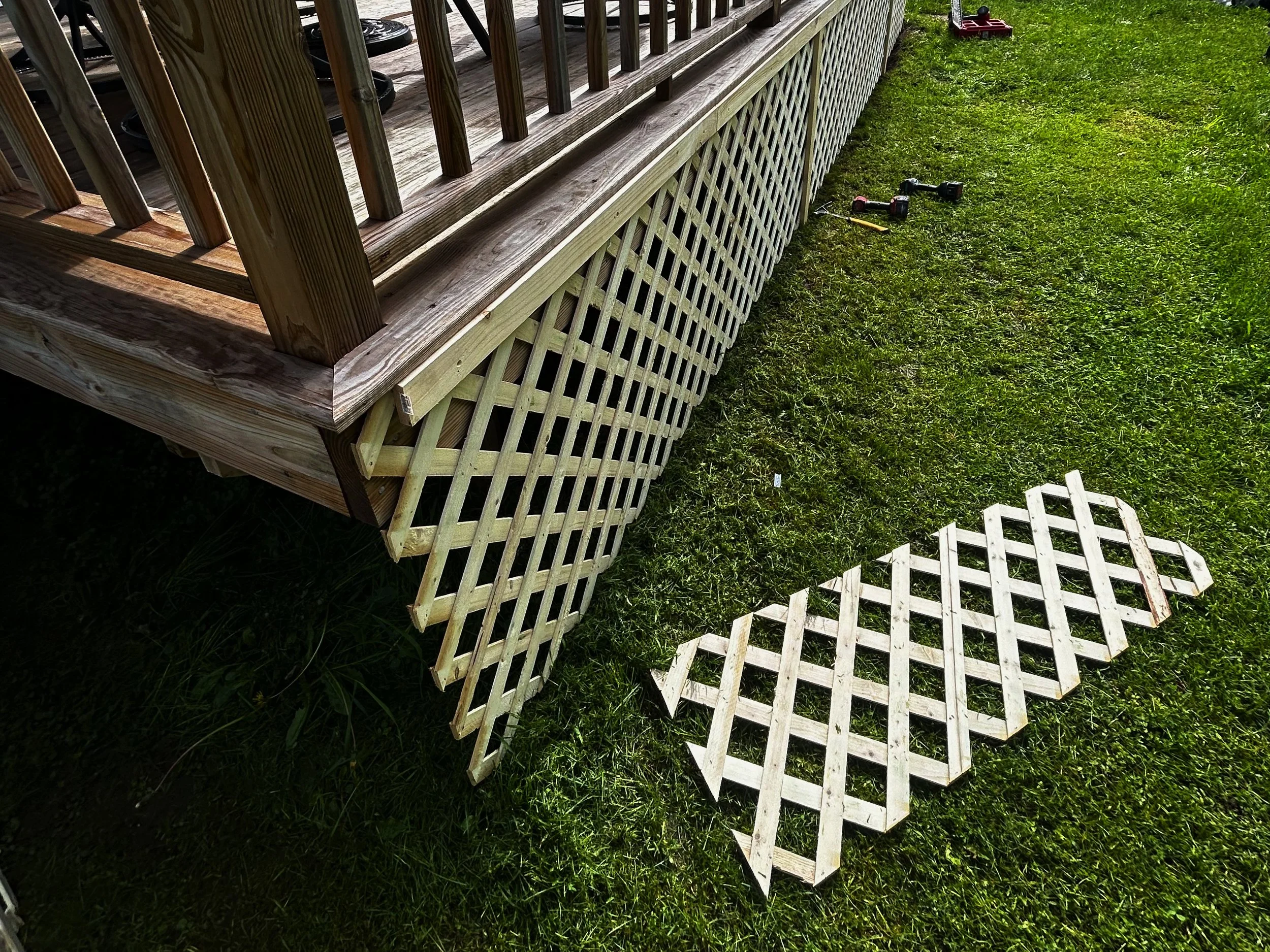 Unassembled lattice panel laying on grass near the base of a wooden deck, with tools and another assembled lattice nearby.