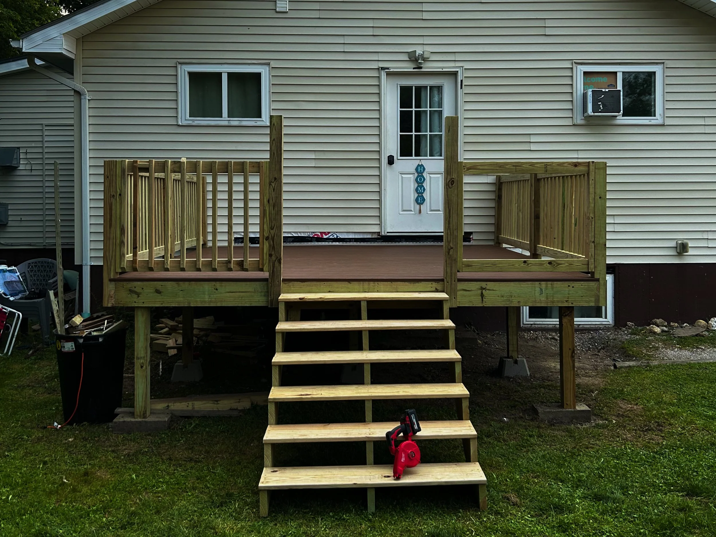 Newly built wooden deck with stairs attached to the back of a house, featuring a railing on three sides and a door leading into the house. Construction tools are visible near the stairs.