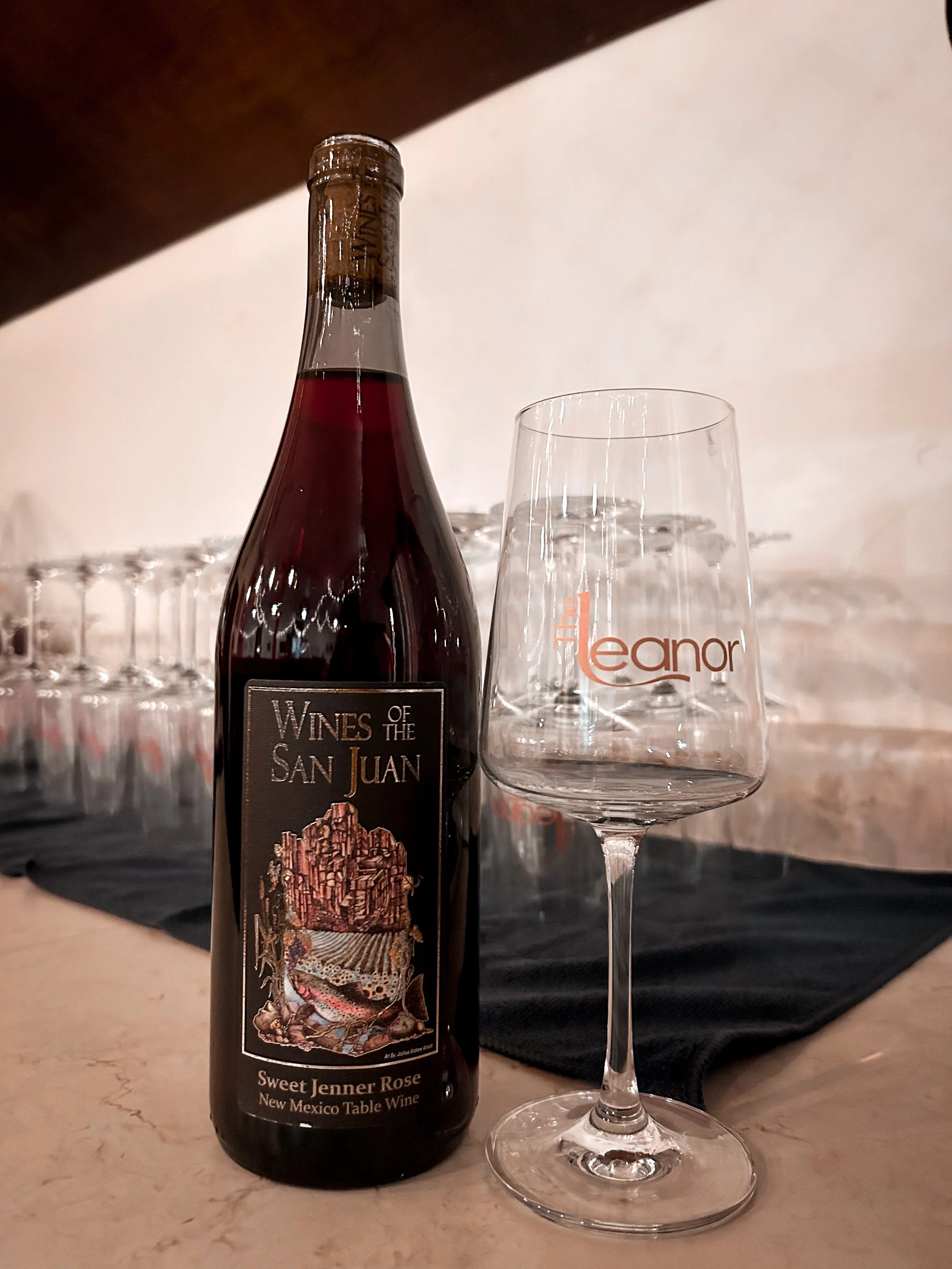 A bottle of Wines of the San Juan Sweet Jenner Rosé wine from New Mexico and a wine glass with the Leanor logo on it on a marble surface in front of a black tablecloth and several empty wine glasses in the background.