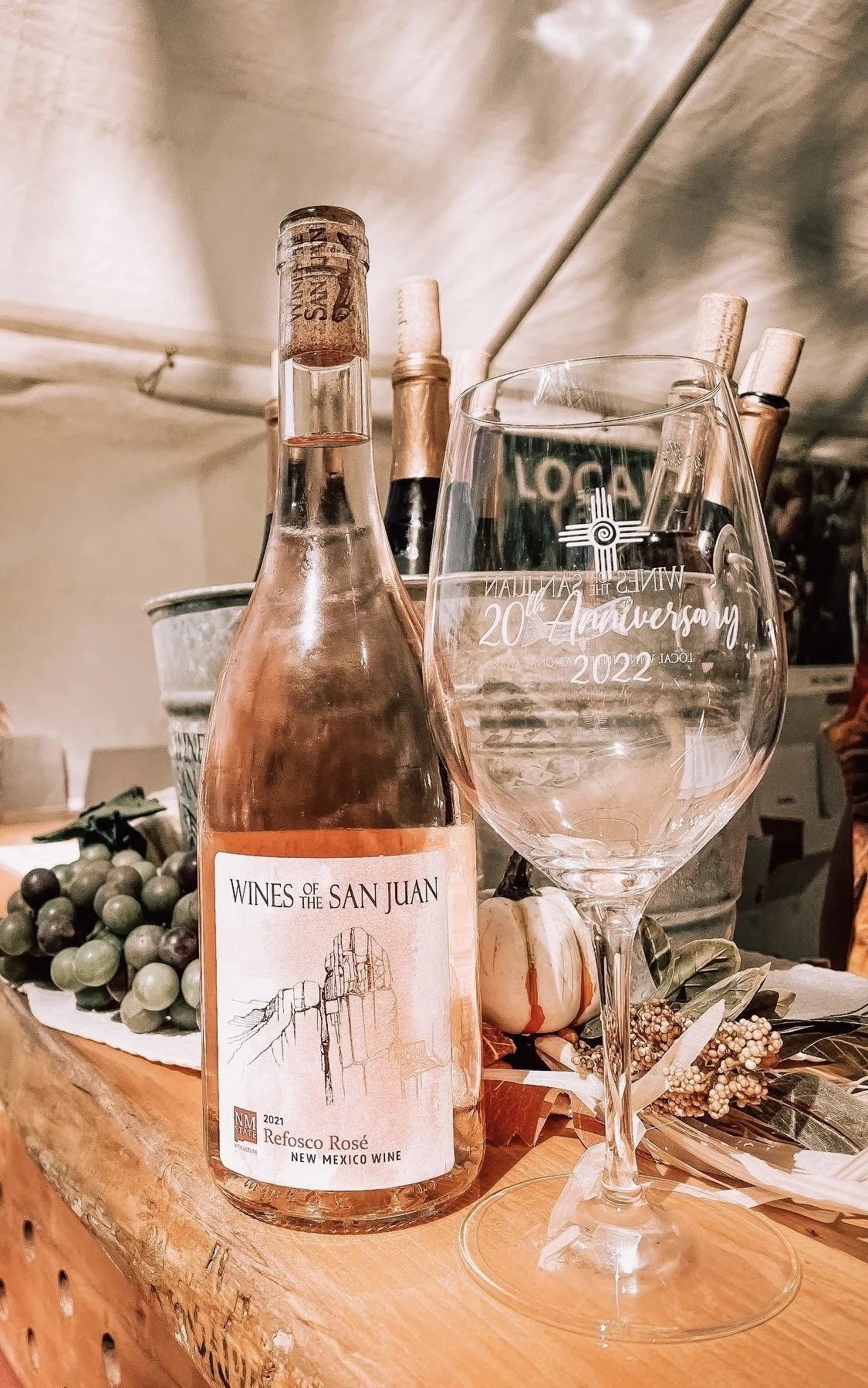 A bottle of Refosco Rosé wine, a wine glass, grapes, a white pumpkin, and decorative dried flowers on a wooden surface, with a background of a beige wall and ceiling.