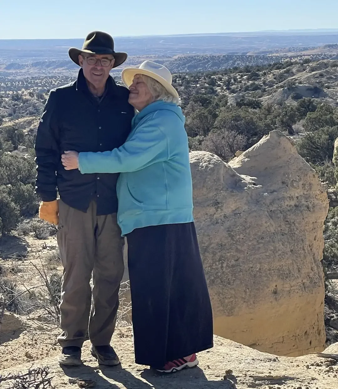 A man and woman embracing outdoors in a rocky, desert landscape with sparse bushes and mountains in the background, both wearing hats and warm clothing.