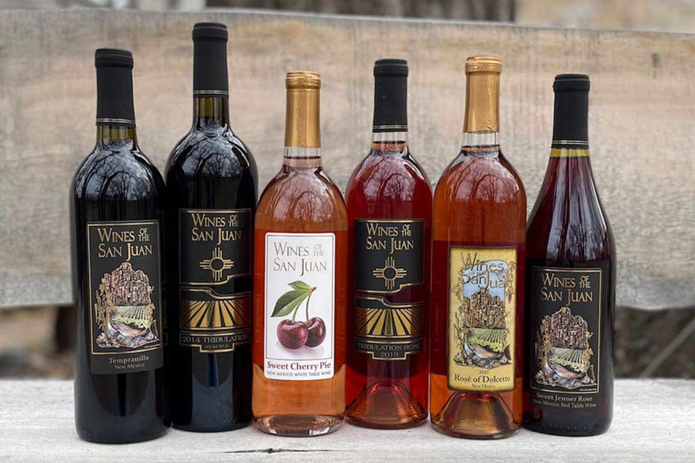 Six bottles of wine, including Tempranillo, Tribulation Reserve, Sweet Cherry Pie, Tribulation Rosé, Rosé of Dolcetto, and Sweet Jenner Rosé, arranged on a wooden surface outdoors.
