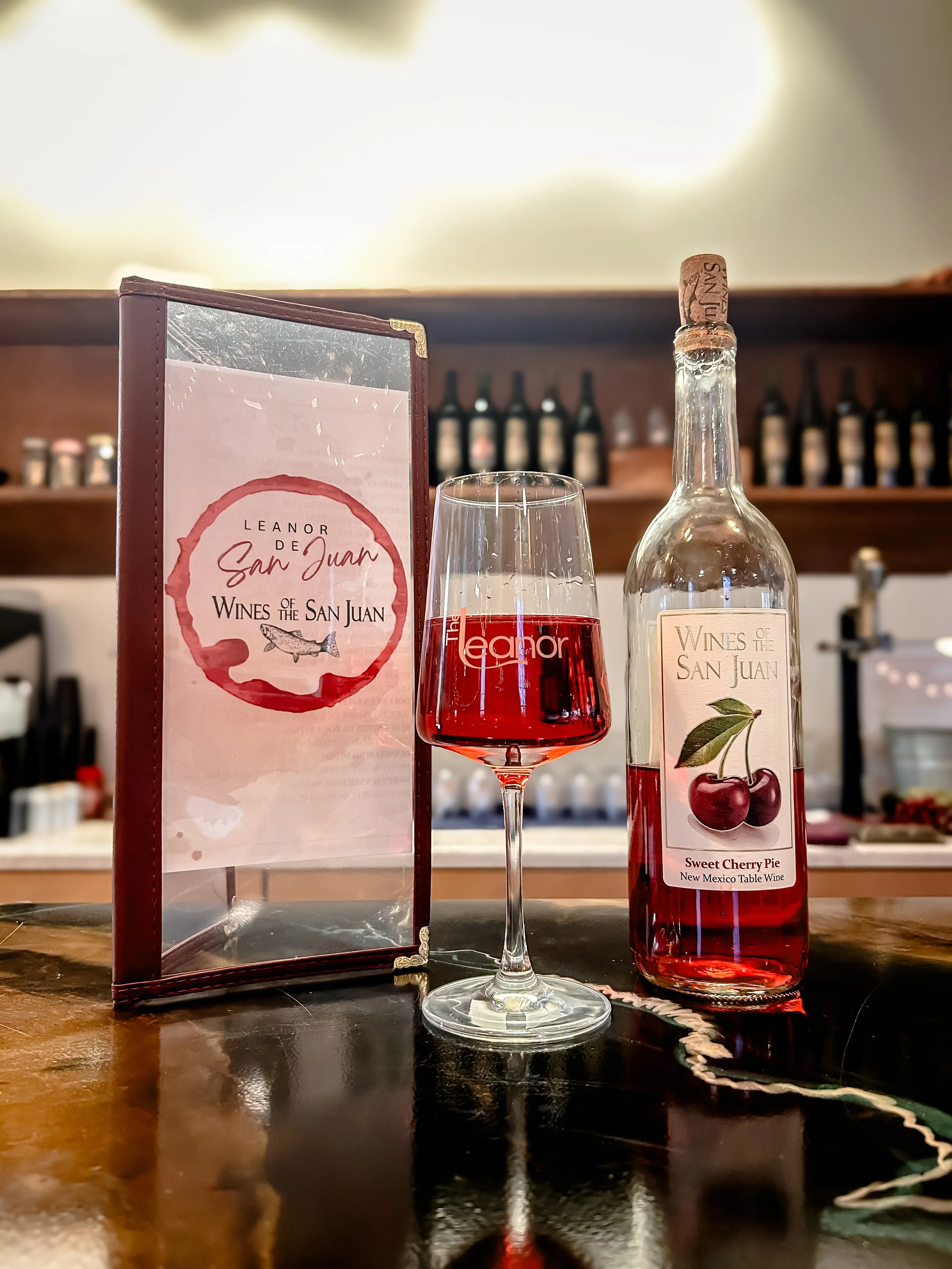 A glass of red wine, a wine bottle with a cherry label, and a menu stand on a wooden bar counter in a wine bar.
