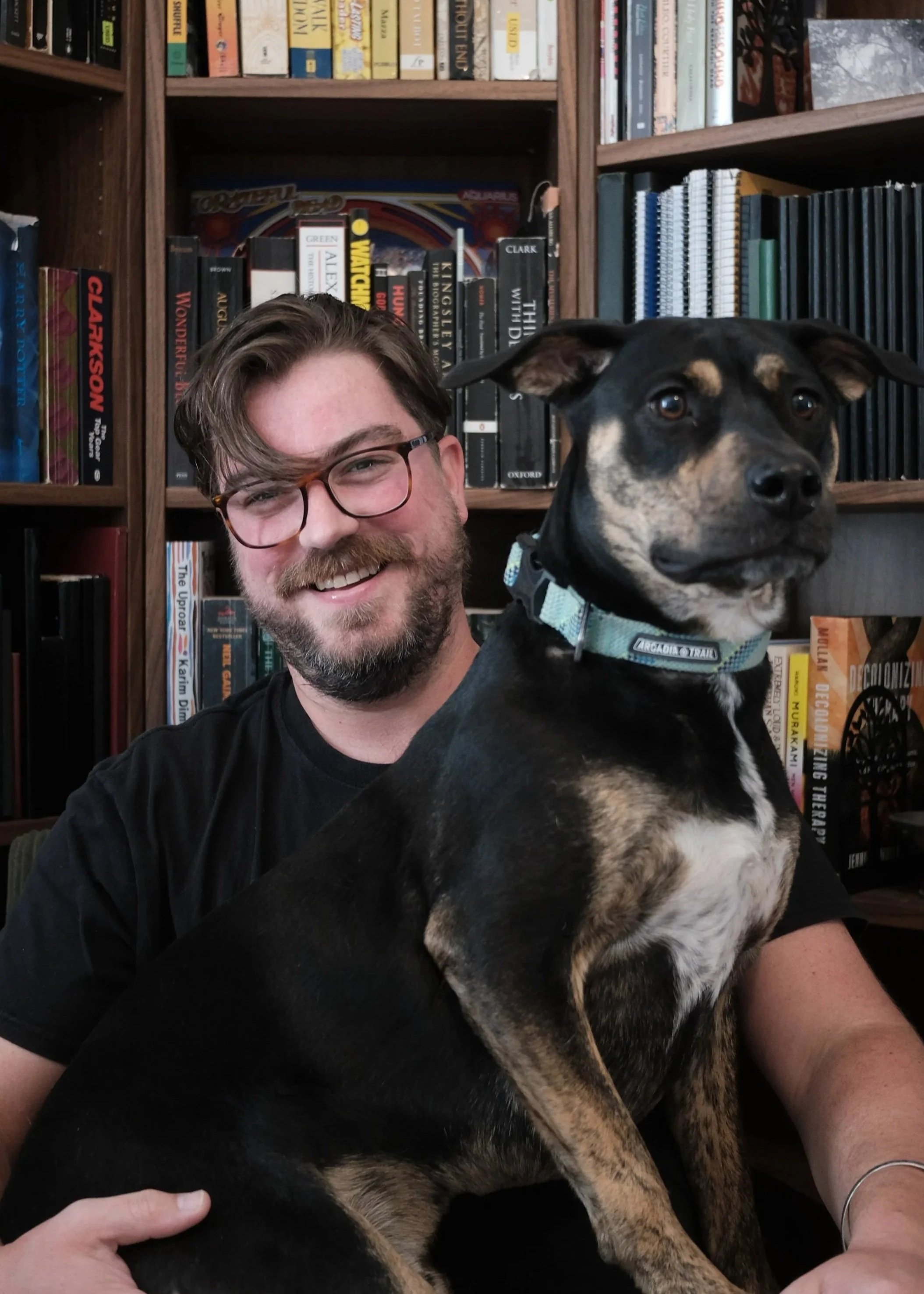 A man with glasses and a beard smiling, sitting in front of a bookshelf, holding a large black and tan dog with a light blue collar.