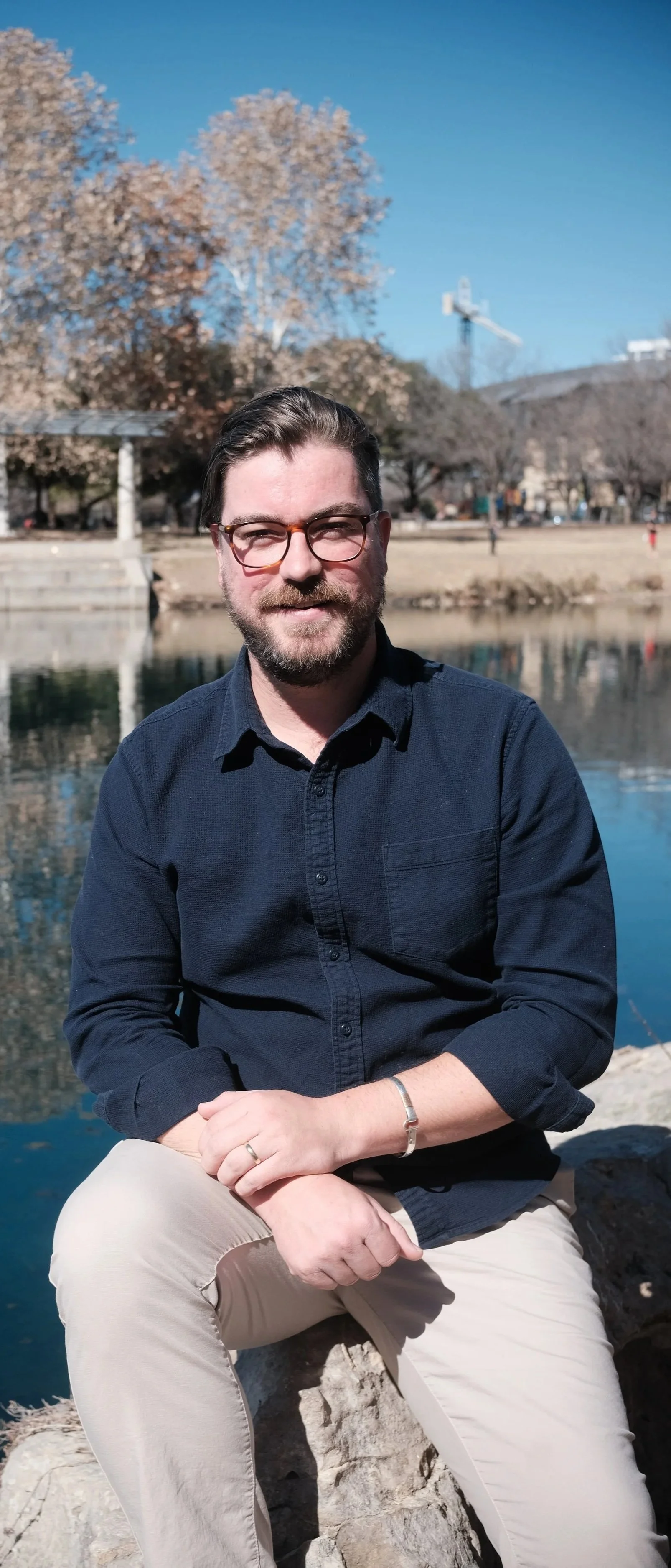 A man with glasses, a beard, and dark hair, wearing a dark shirt and light-colored pants, sitting on a rock beside a river with trees and a building in the background during sunny weather.