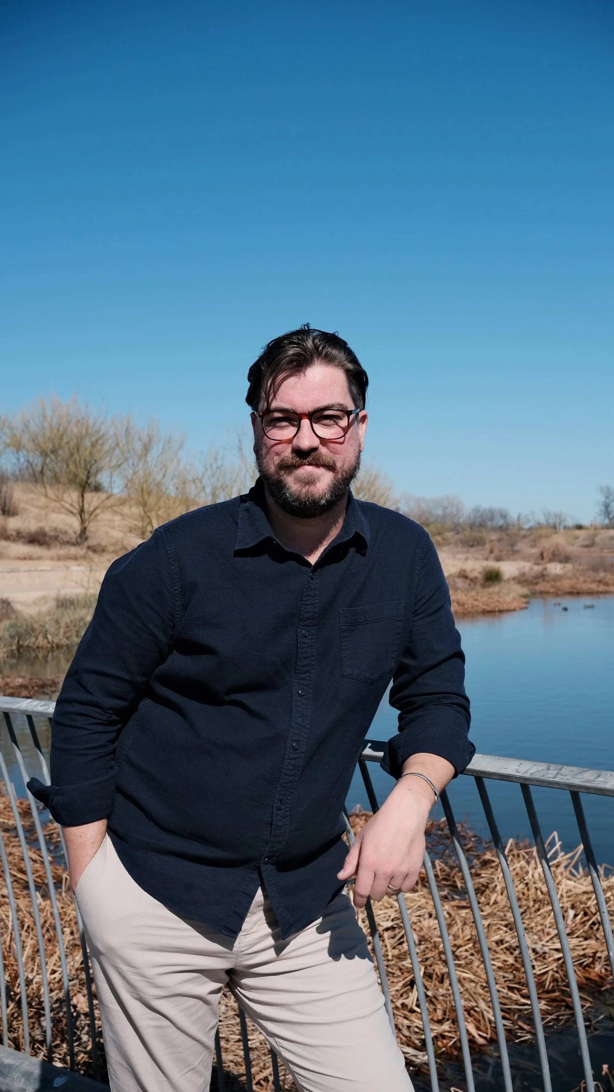 A man with glasses, dark hair, and a beard, wearing a navy button-up shirt and beige pants, leaning on a railing near a body of water with trees and bushes in the background under a clear blue sky.
