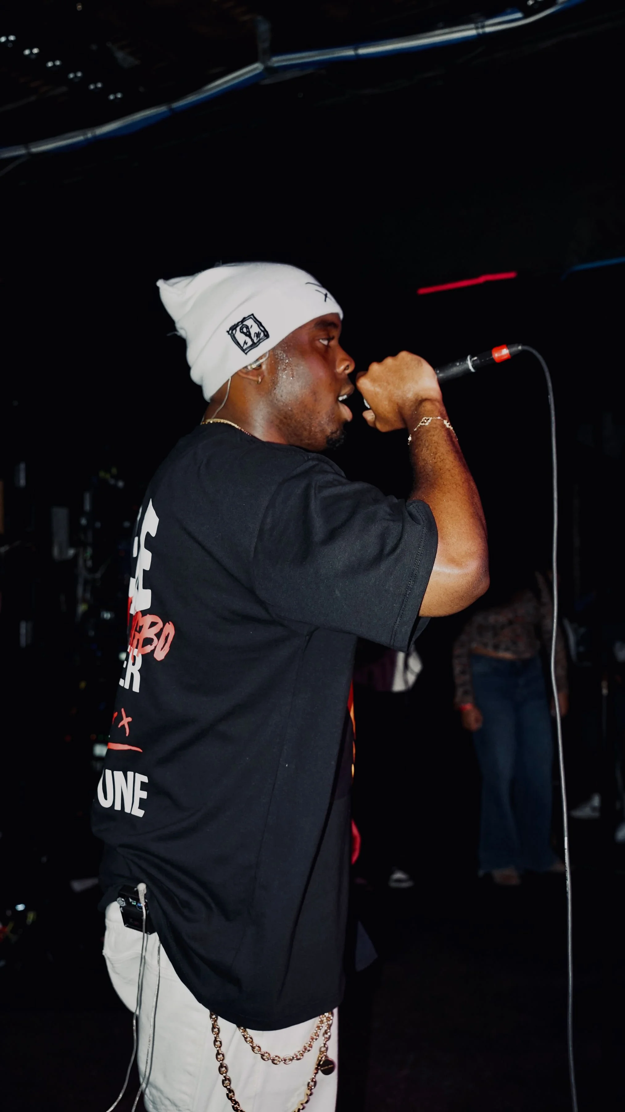 A man wearing a white beanie and black T-shirt singing into a microphone at a dark indoor venue.
