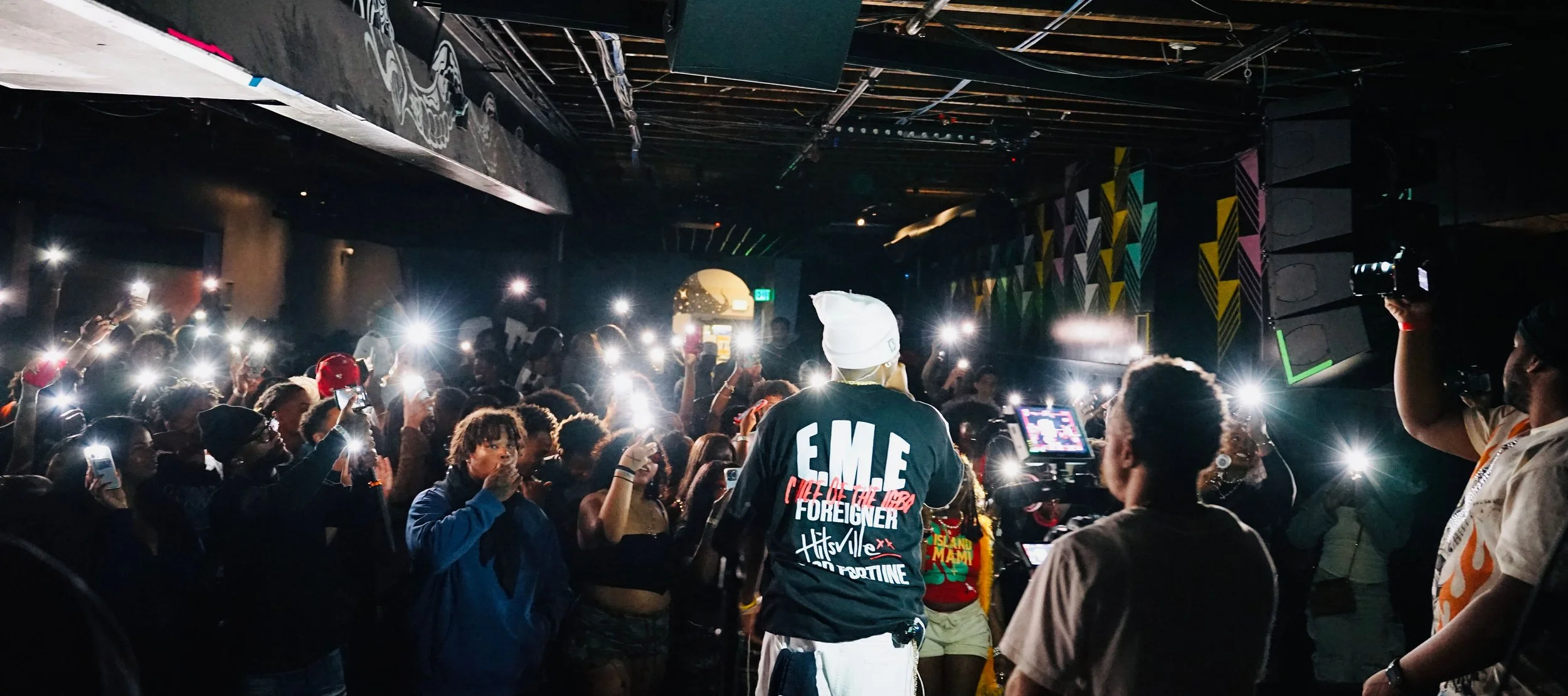 Crowd attending a music concert or event in a dark indoor venue, many people holding up smartphones with flashlights on, focusing on a person in the foreground wearing a black T-shirt and white beanie, with colorful graffiti-style designs on the wall