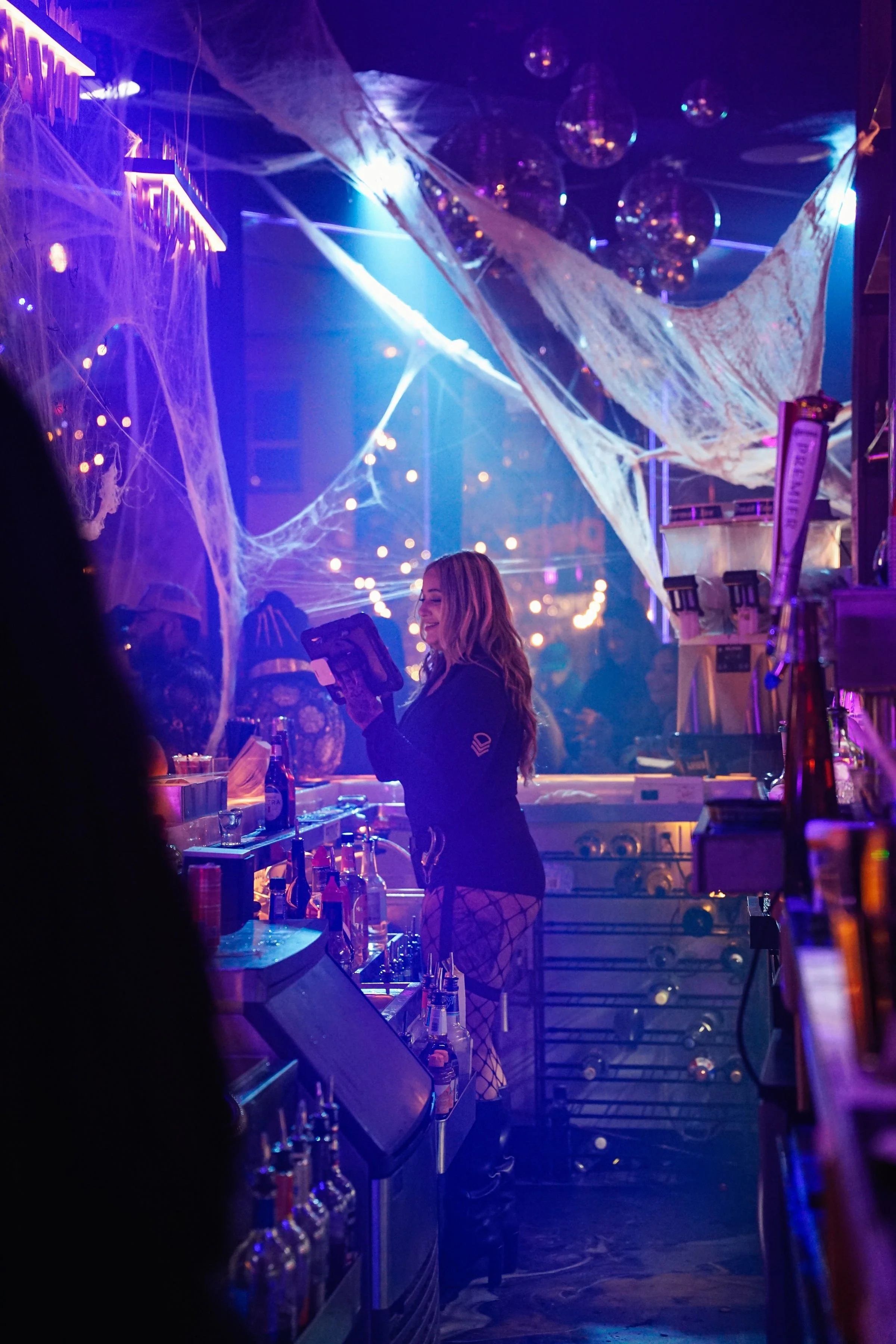 A woman working behind a bar in a nightclub decorated with Halloween-themed decorations like fake spider webs and hanging ornaments, illuminated with purple and blue neon lights.