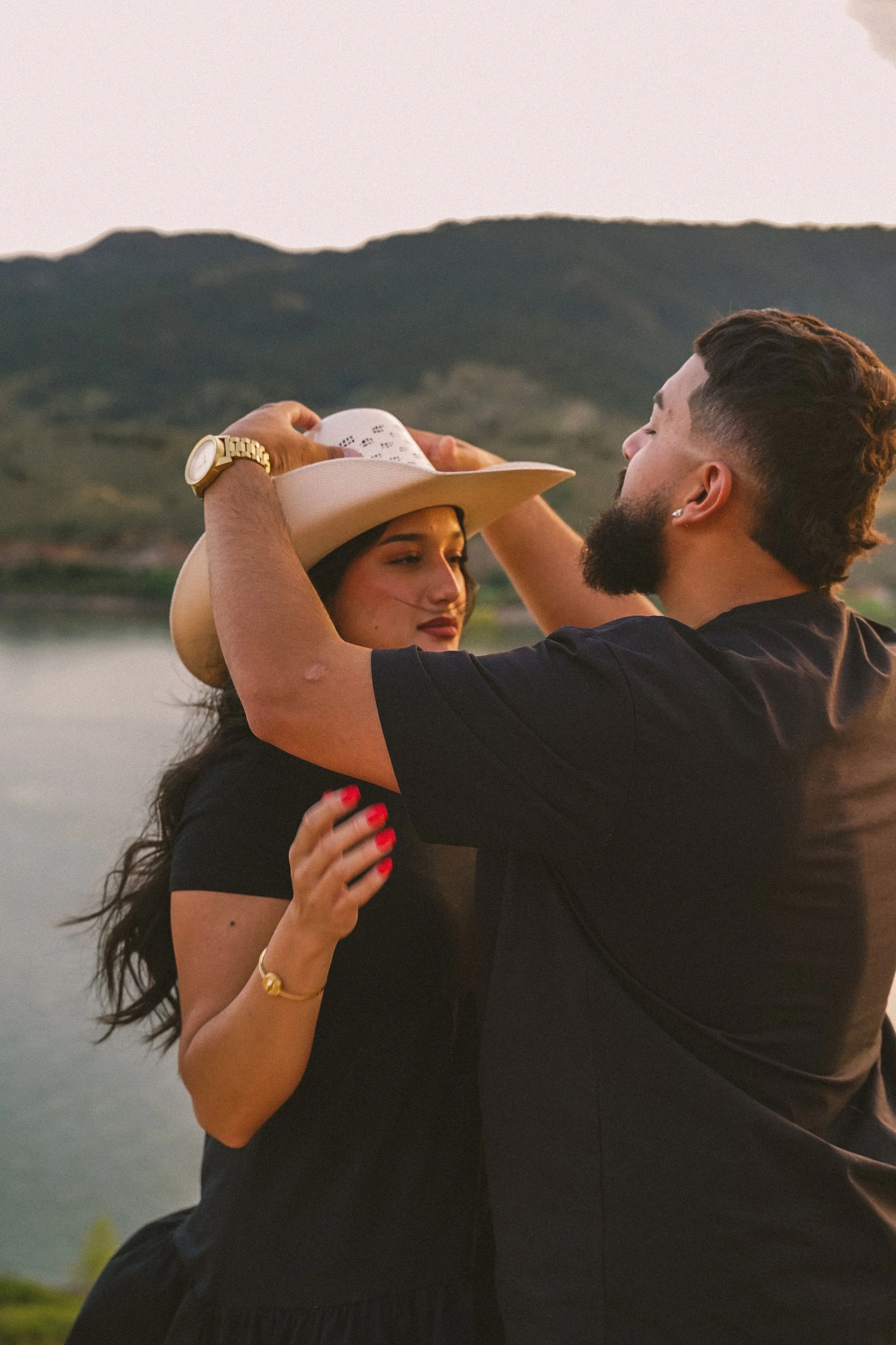A couple dancing outdoors by a lake during sunset, with mountains in the background. The woman wears a hat, black shirt, and gold jewelry, while the man has a beard and is dressed in black.