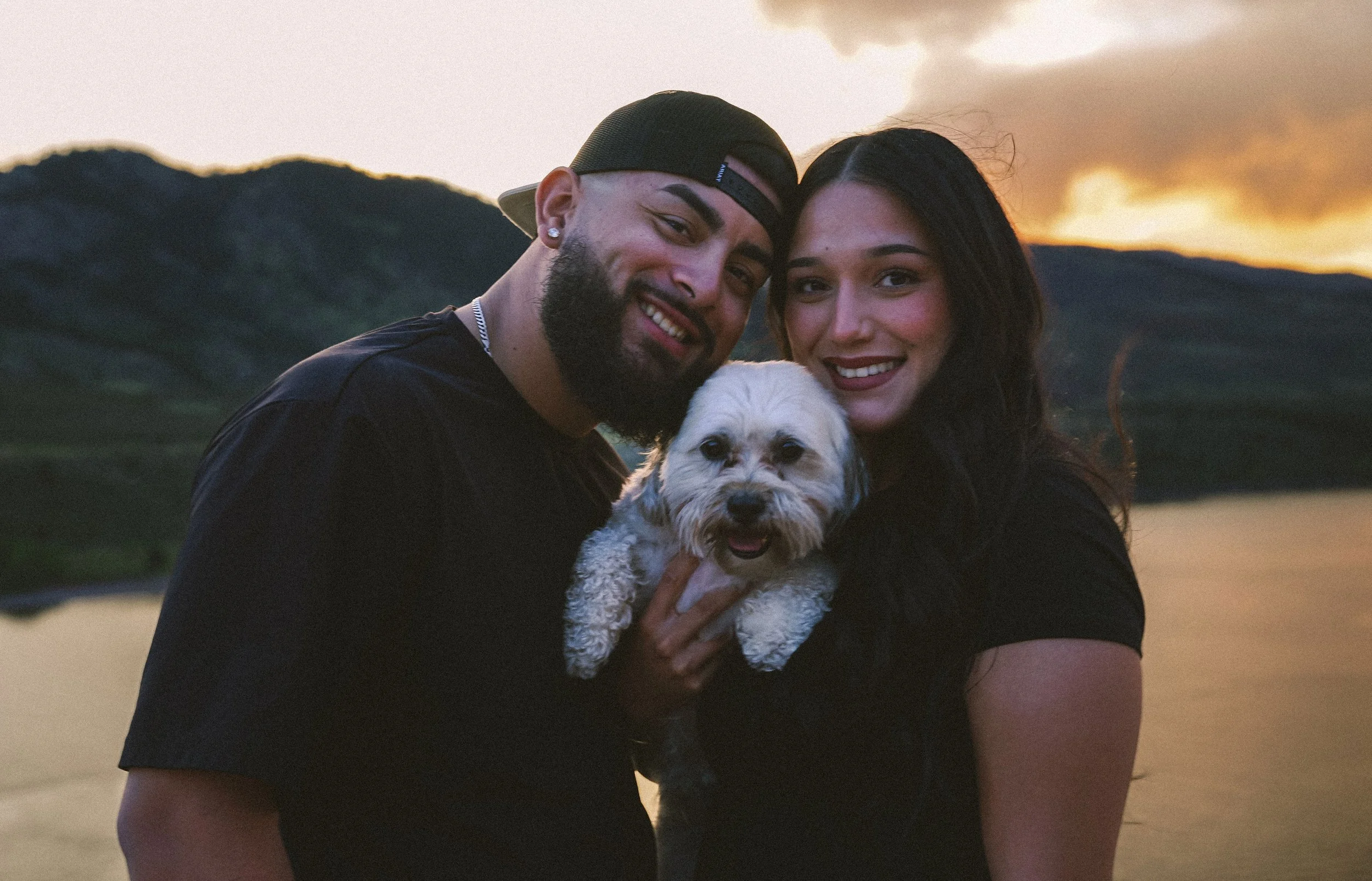 A happy couple and their small dog enjoying sunset by the water with mountains in the background.