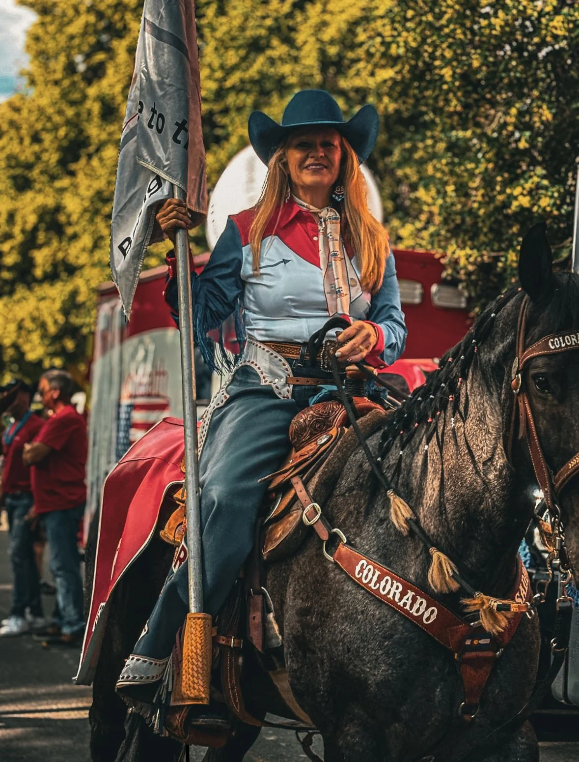 Woman dressed as a cowgirl, wearing a cowboy hat and Western attire, riding a horse during a parade or event with people and trees in the background.