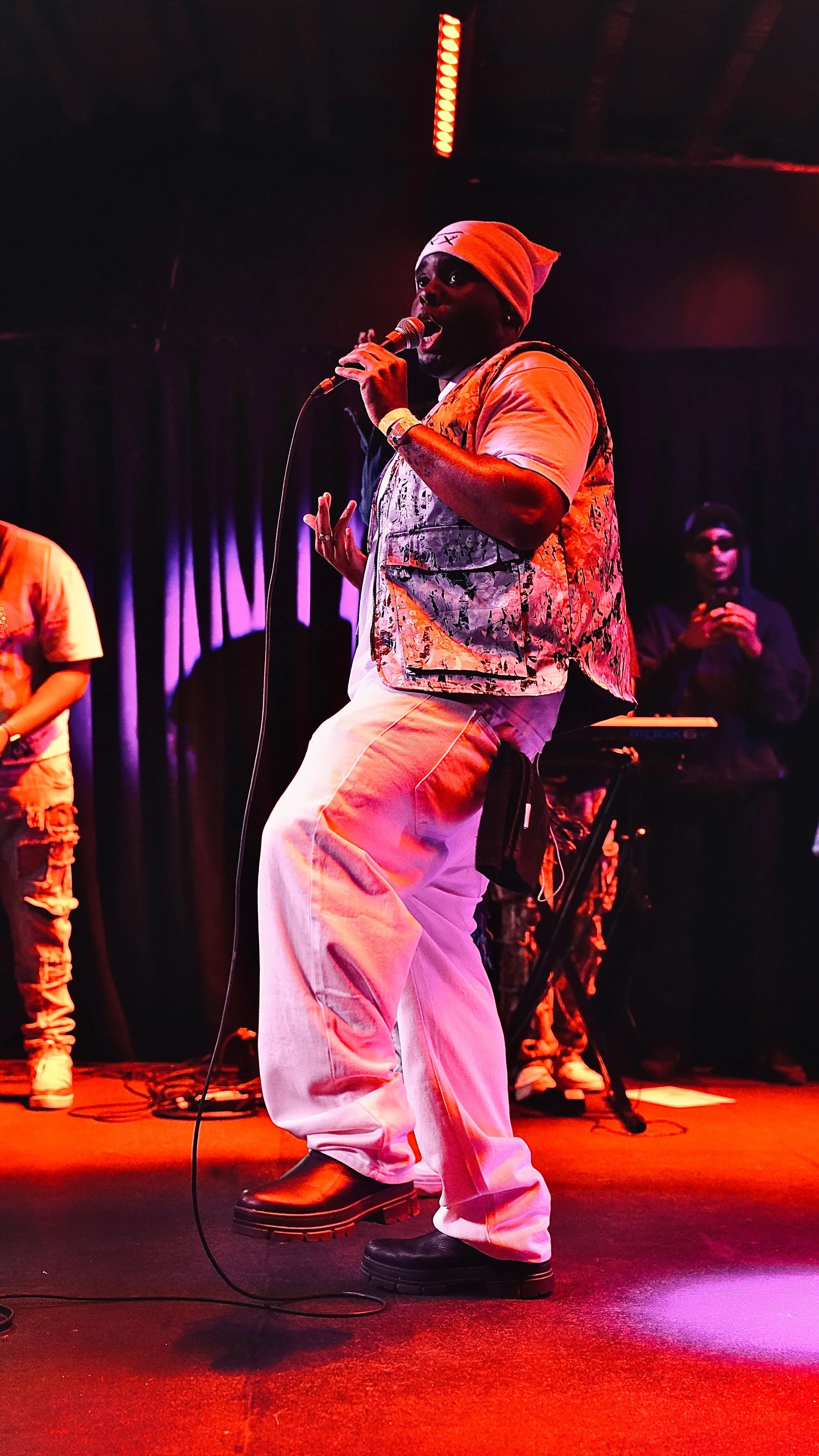 A singer performing on stage with a microphone, wearing a beanie, a patterned vest, light-colored pants, and black boots, with a dark curtain backdrop and stage lighting.