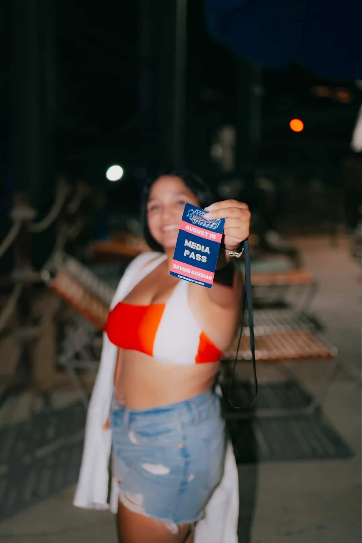 Woman holding a media pass at night, wearing a red and white top and denim shorts, smiling towards the camera.