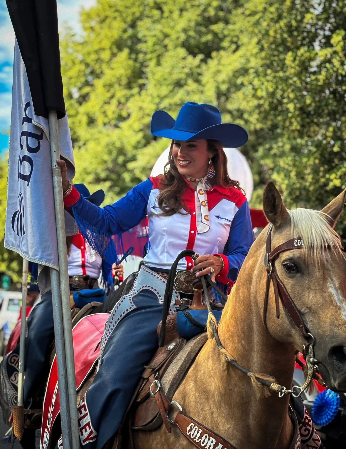 A woman dressed in Western attire, wearing a large blue cowboy hat and a red, white, and blue shirt, sits on a light brown horse. She is smiling and holding a flagpole, and there are trees in the background.
