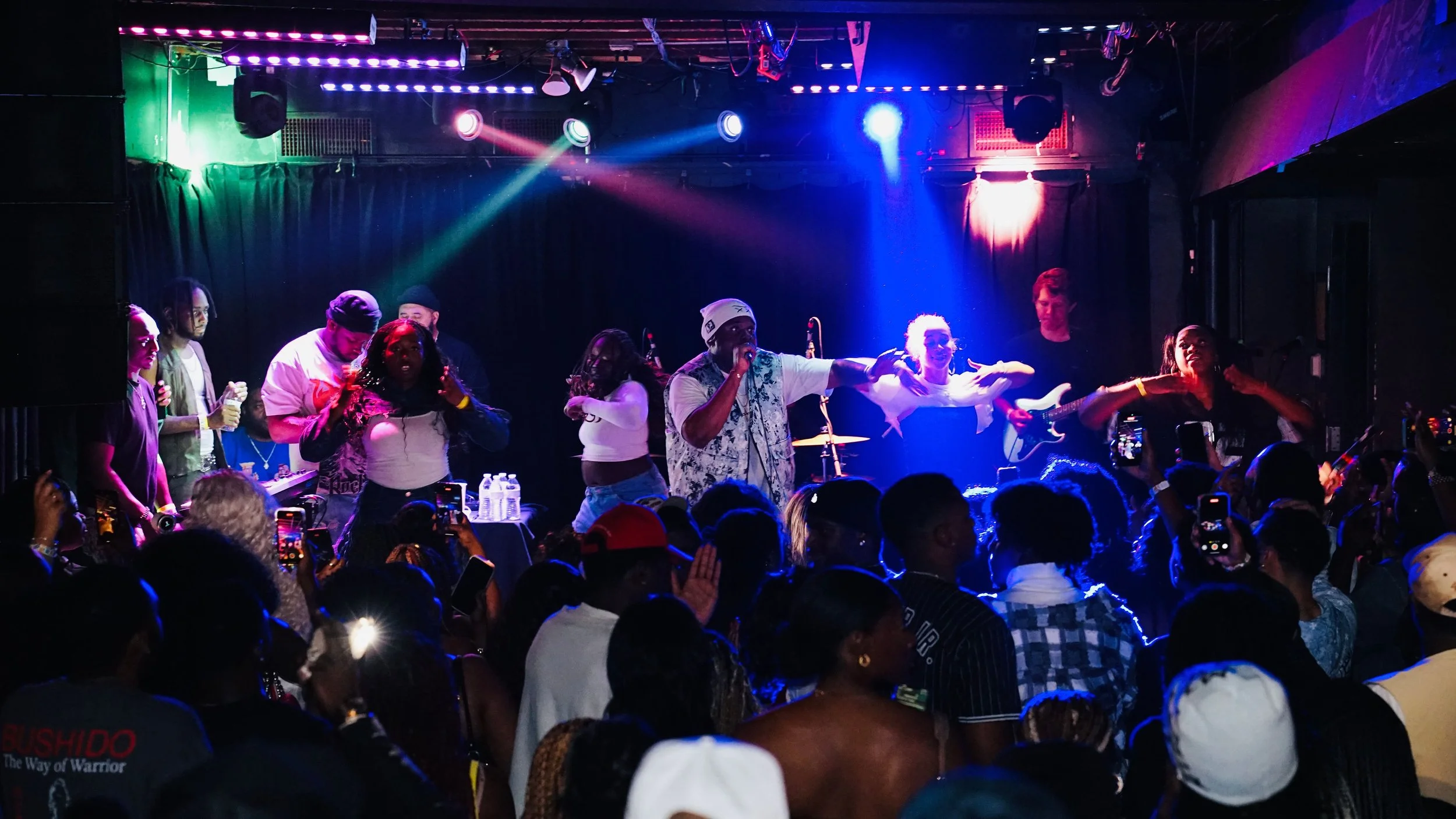 Performer on stage with backup singers and musicians during a concert, audience taking photos, colorful stage lighting.