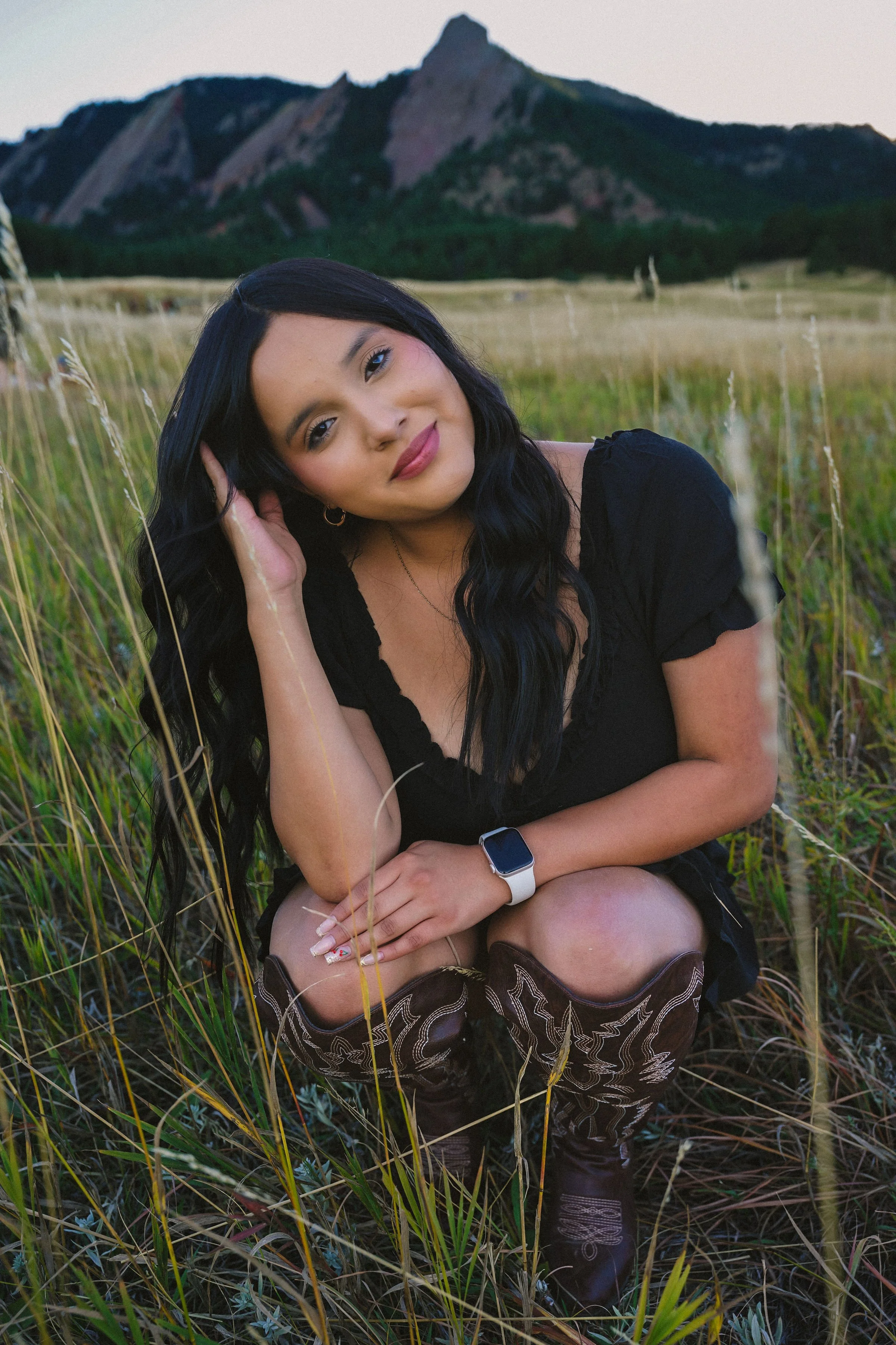 A woman with long black hair and a smartwatch on her wrist is sitting in a grassy field with mountains in the background.
