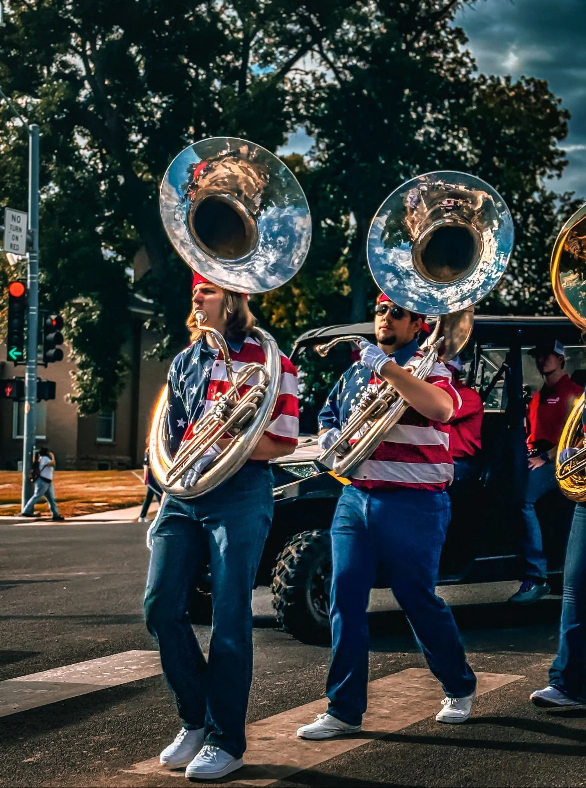 Two musicians in a parade, dressed in American flag-themed shirts, playing sousaphones and marching across a crosswalk. Others are visible in the background, with trees and traffic signals.