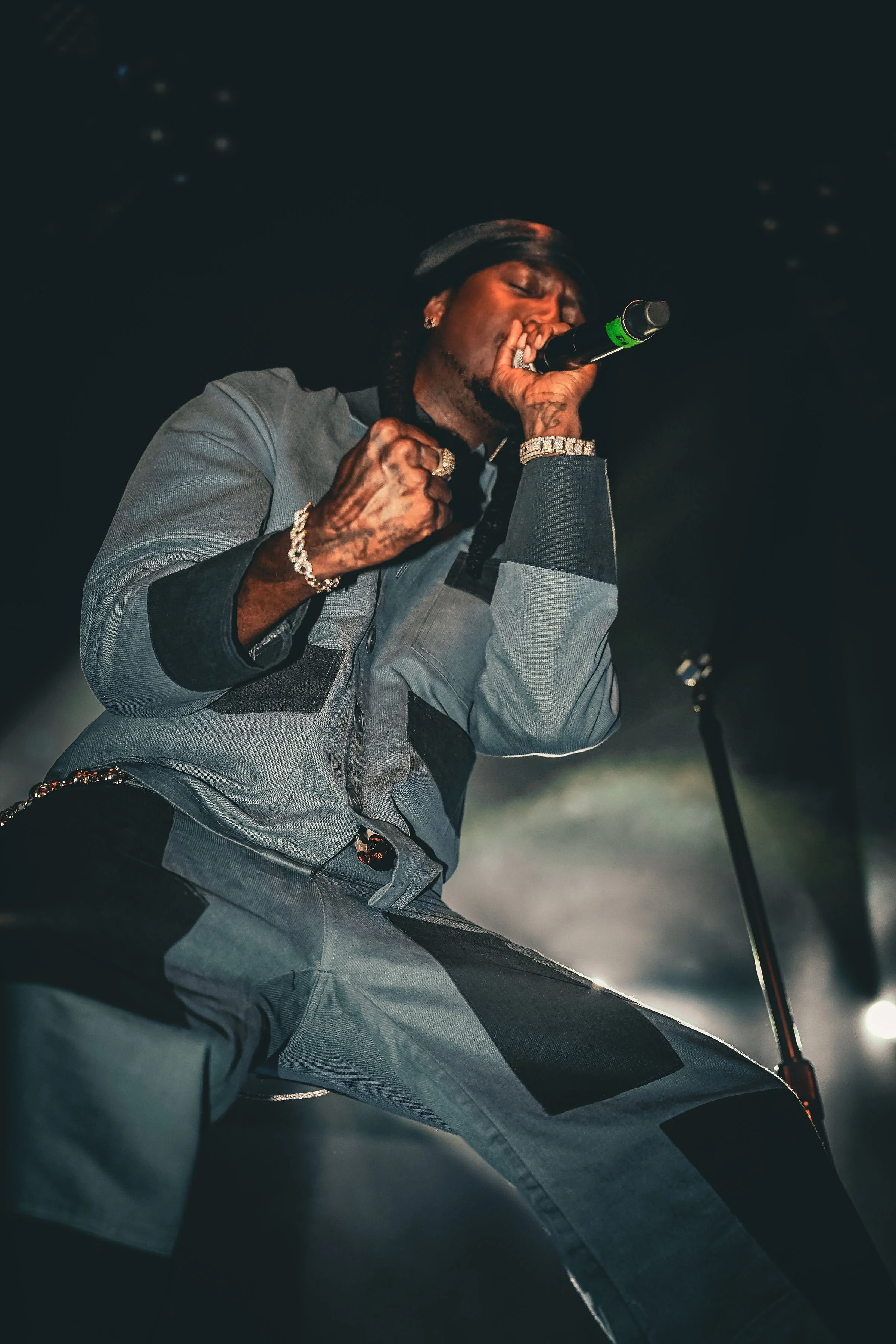 A male singer performs passionately on stage, holding a microphone and raising his fist, wearing a gray and black outfit with jewelry, under dark stage lighting.