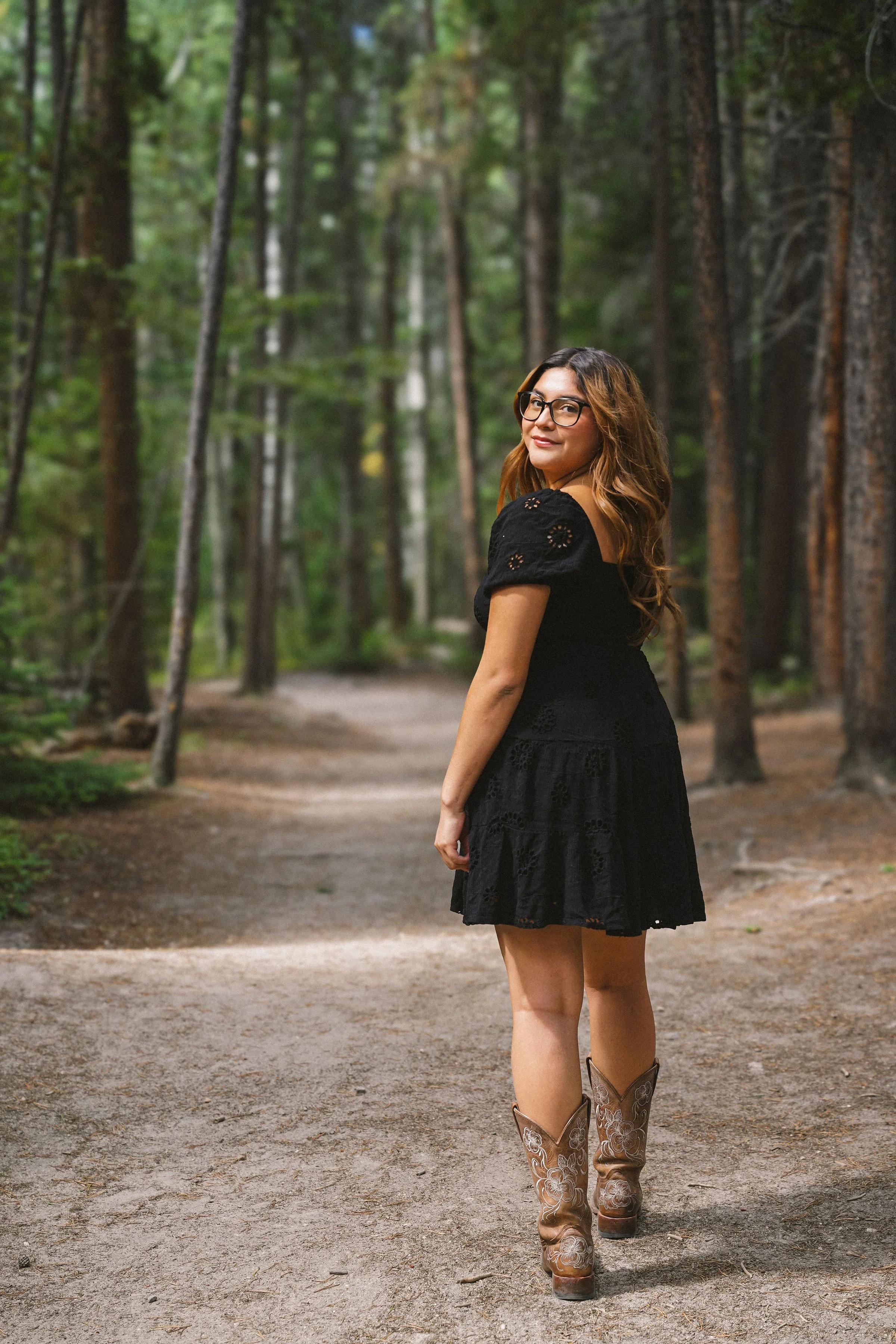 A woman with long wavy hair and glasses walking in a forest, wearing a black dress and cowboy boots.