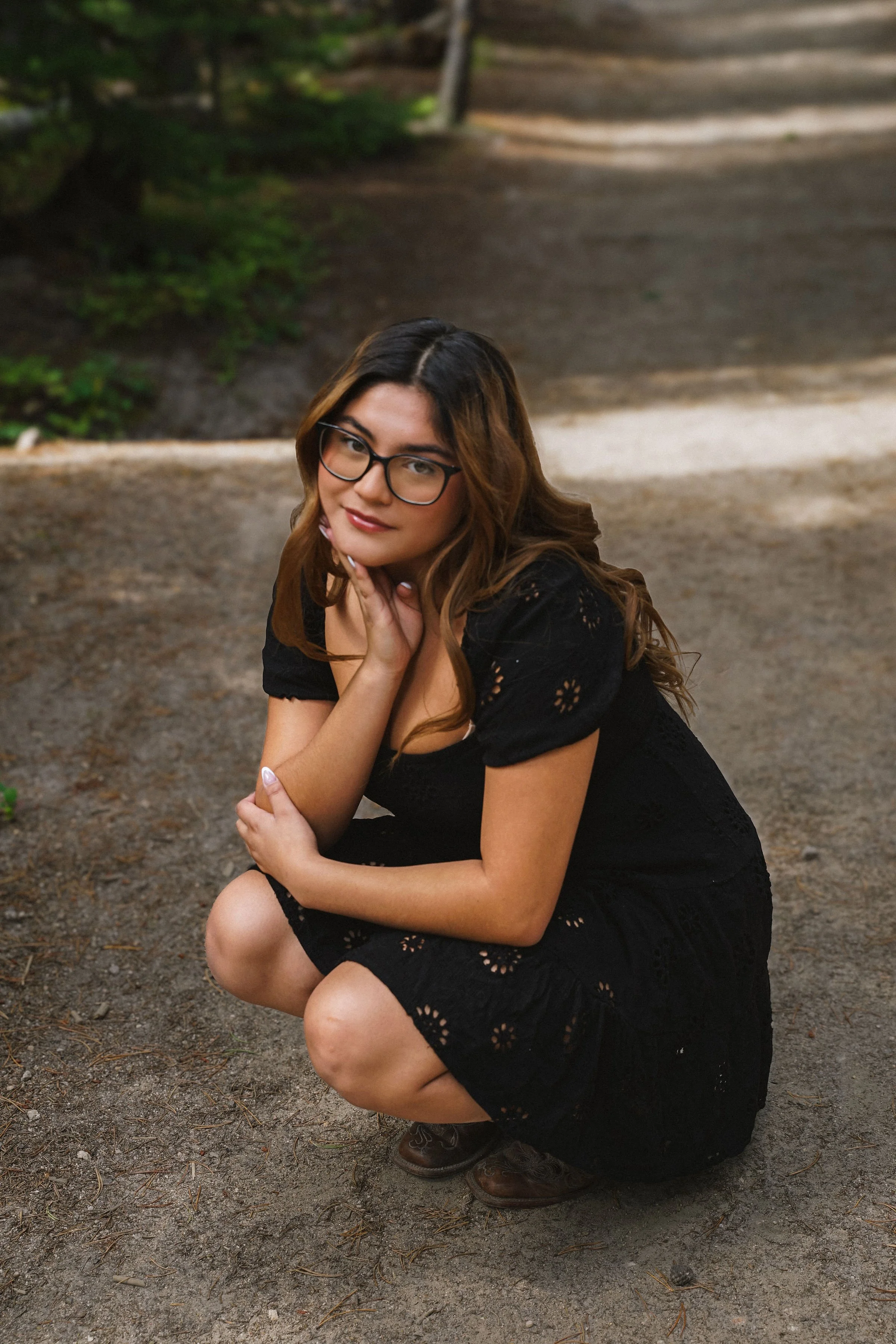 A woman with glasses and long brown hair squats on a dirt path in a park, wearing a black dress and brown shoes, with greenery and trees in the background.