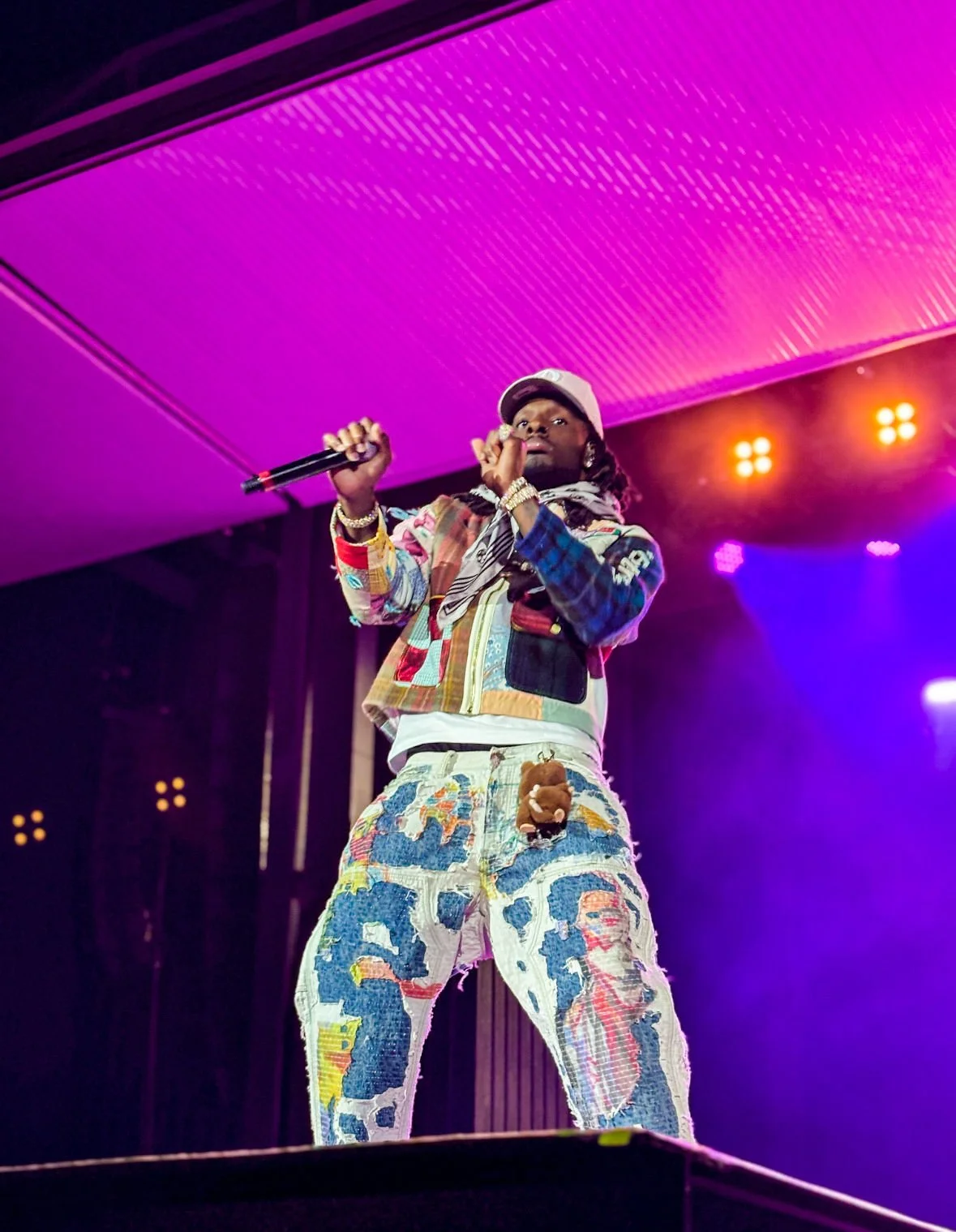 A rapper on stage holding a microphone, wearing a colorful patchwork jacket, distressed denim pants with patches, and a baseball cap, with purple and orange stage lighting in the background.