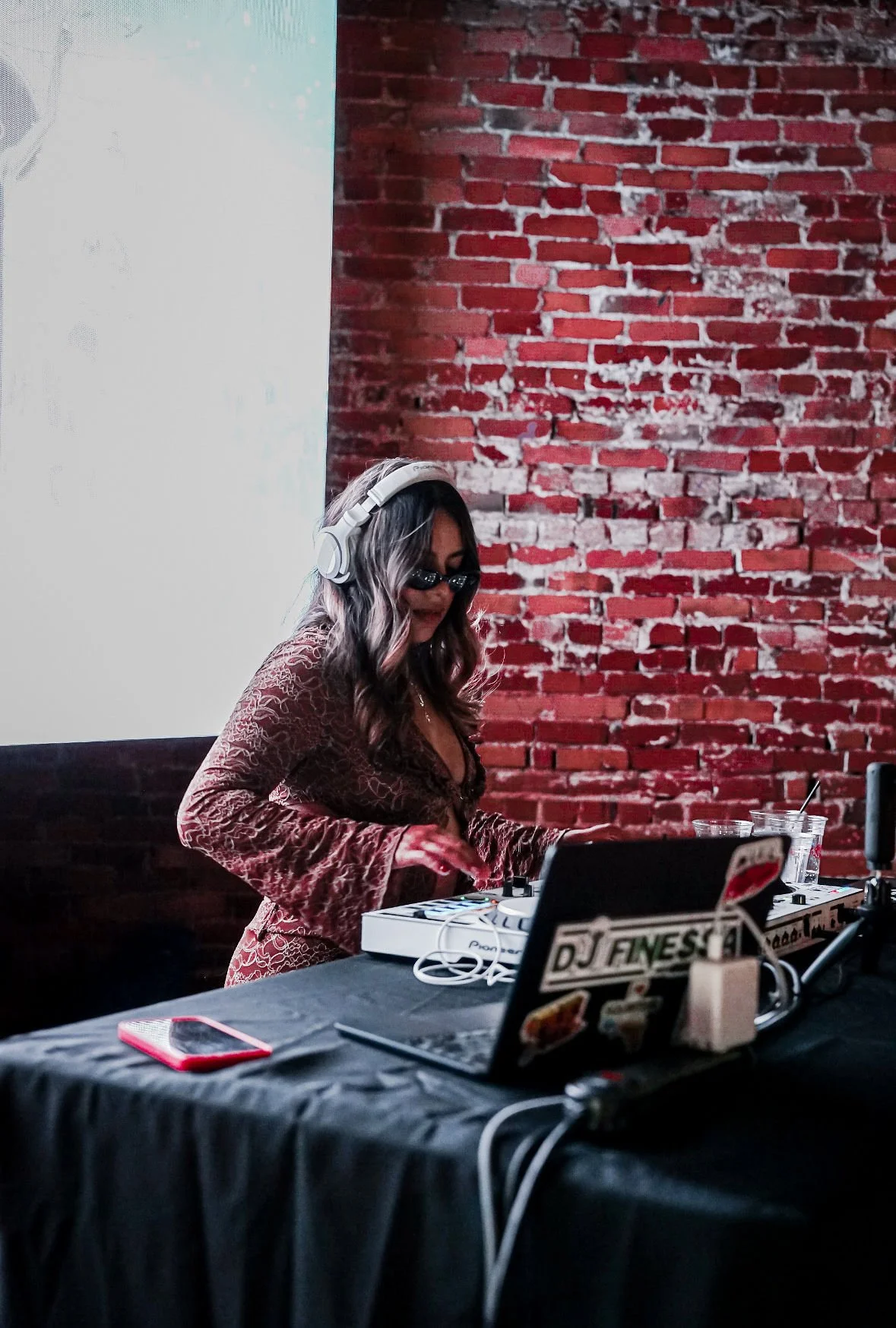 A female DJ with long wavy hair, wearing sunglasses, headphones, and a brown patterned dress, is standing at a DJ setup with a mixer, laptop, and other equipment against a red brick wall.