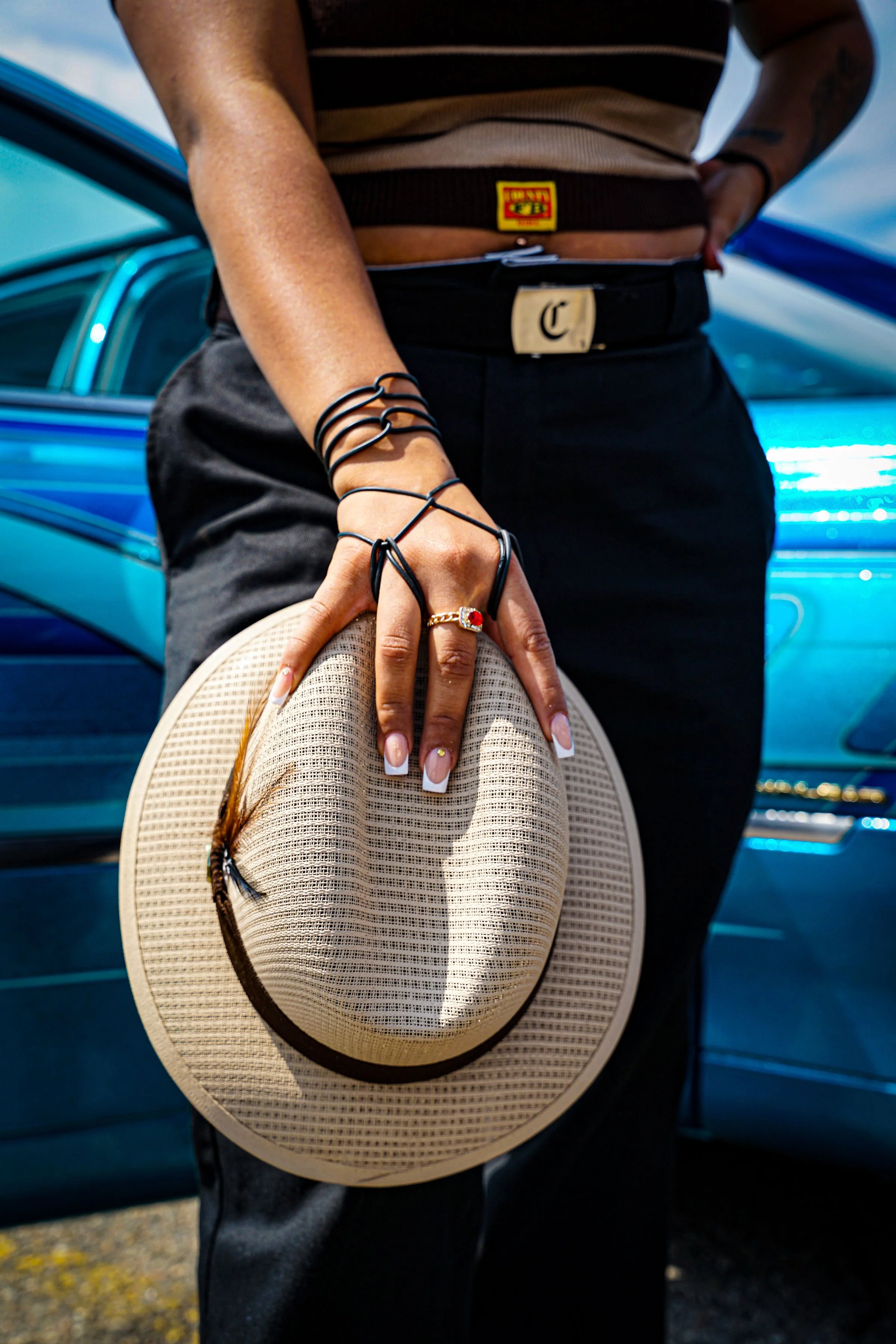 Person holding a beige straw hat with a black band, standing in front of blue cars, wearing black pants, a striped top, and various jewelry including rings and bracelets.