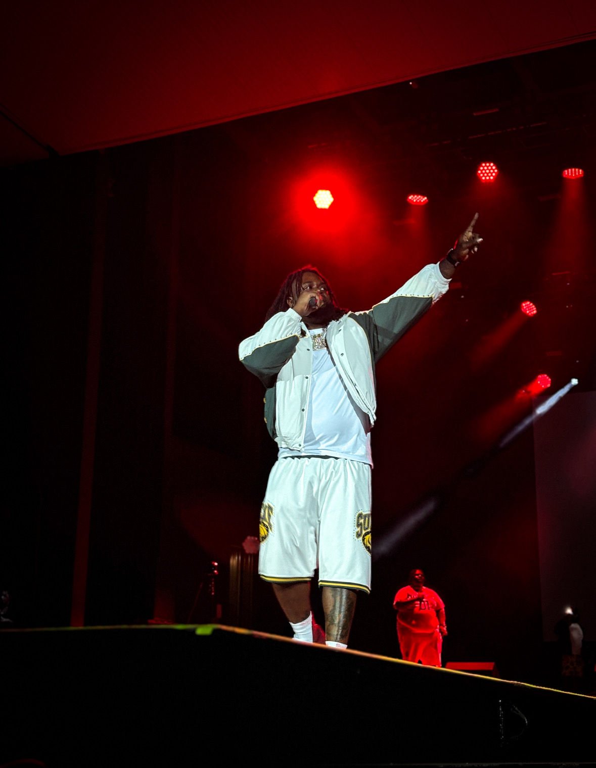 A male performer on stage dressed in sportswear, holding a microphone, pointing upward with one hand, with stage lights shining down, and a person in red standing in the background.