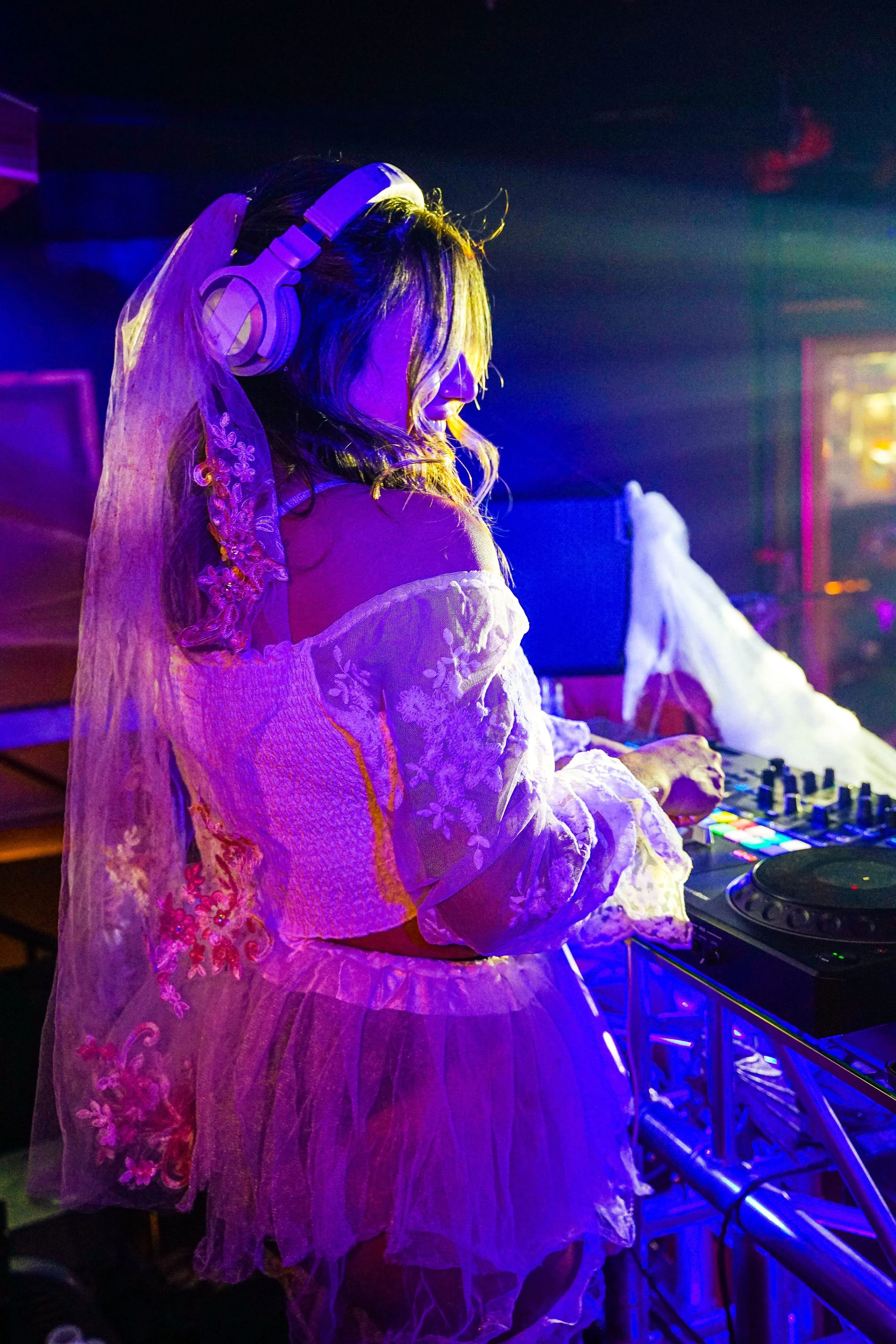 A woman DJ wearing a white off-shoulder dress with embroidered floral patterns and a sheer veil, standing at a DJ booth with colorful LED lights, in a dimly lit nightclub with vibrant lighting.