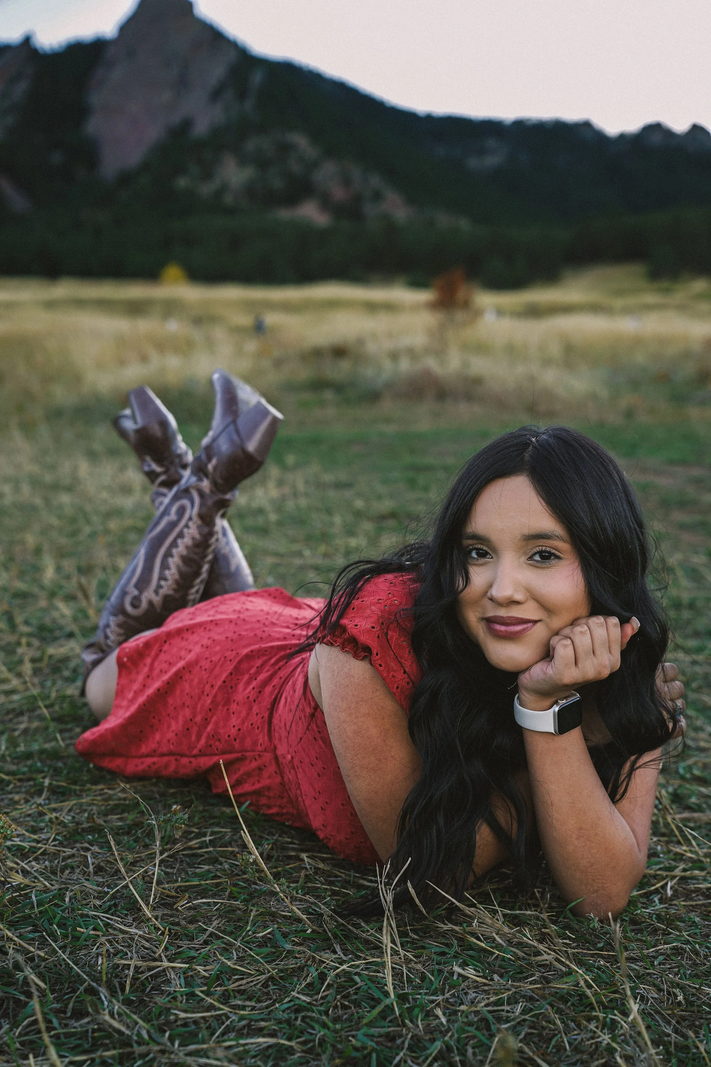 A woman with long dark hair lying on her stomach in a grassy field, resting her chin on her hand, wearing a red dress and patterned boots. Mountains are visible in the background.