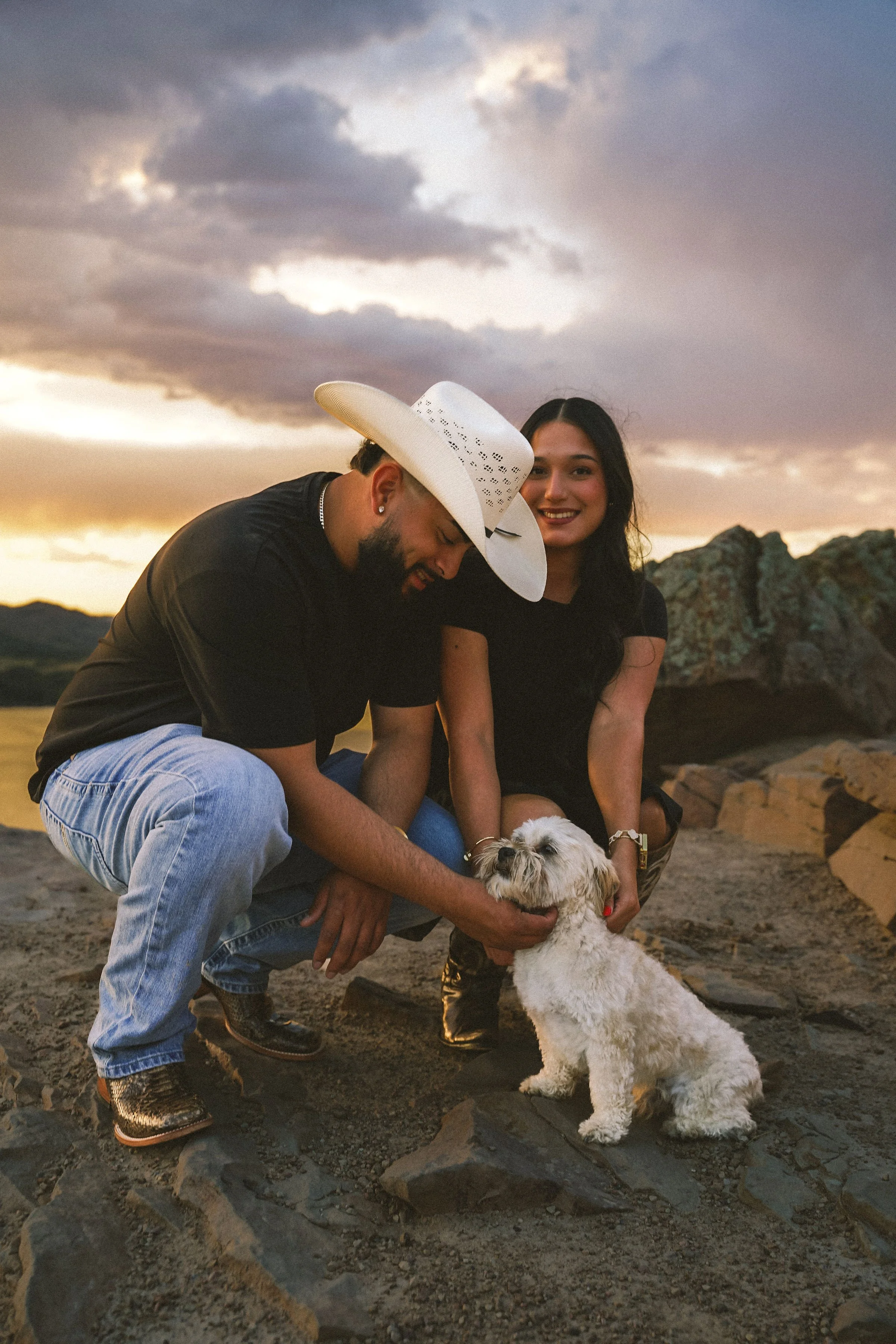 A man in a cowboy hat and a woman with long dark hair smiling as they pet a small white dog on a rocky beach during sunset.