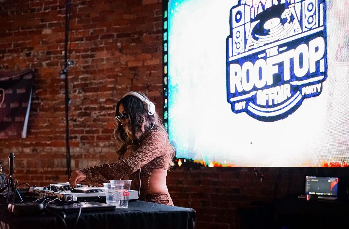 A woman with long wavy hair wearing sunglasses and a brown lace top is DJing at a club or event, standing behind a DJ booth with equipment. There is a large illuminated sign in the background that says 'The Rooftop' with a cityscape graphic, and the 