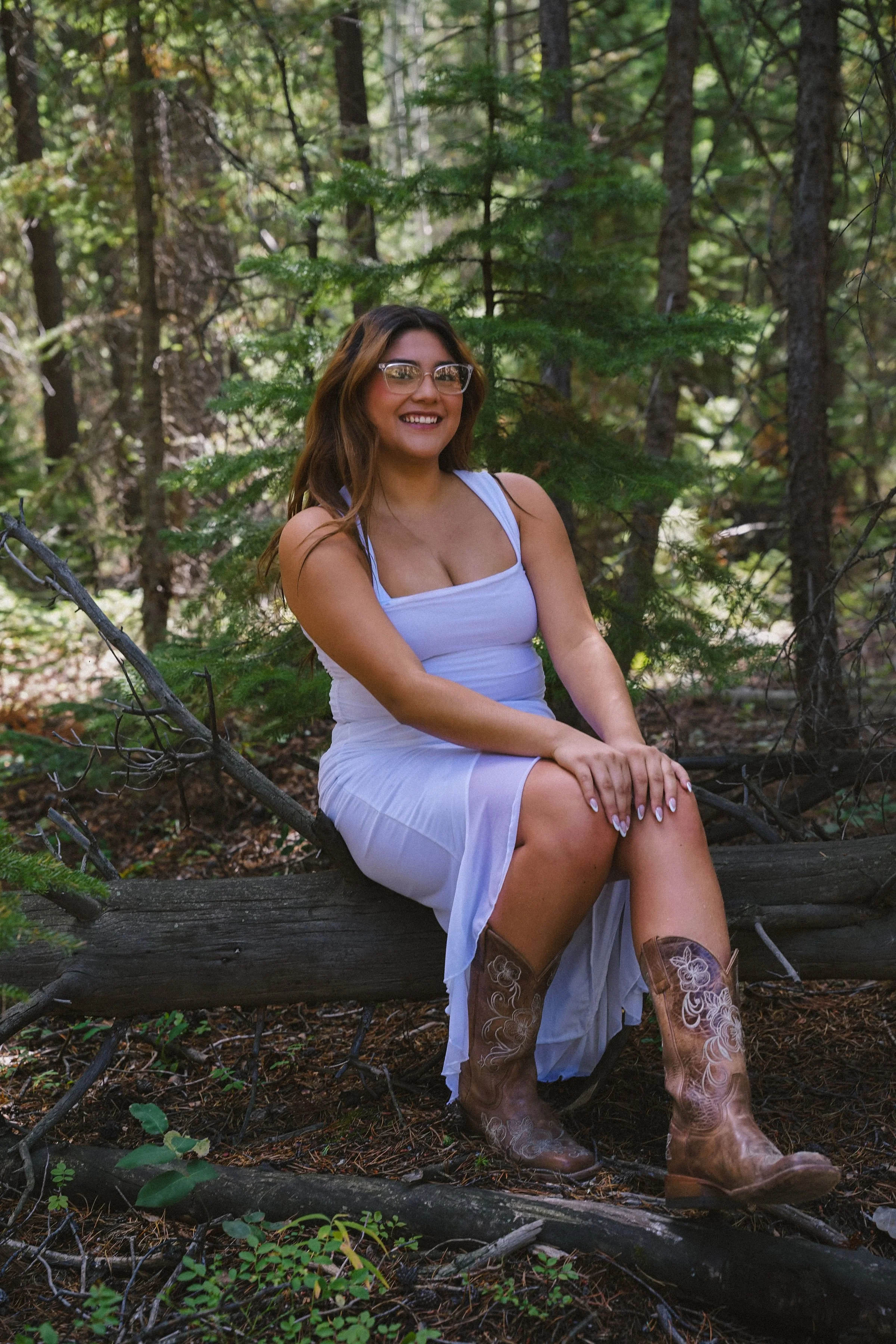 A woman with glasses and long hair, wearing a white dress and cowboy boots, sitting on a fallen log in a wooded area and smiling at the camera.