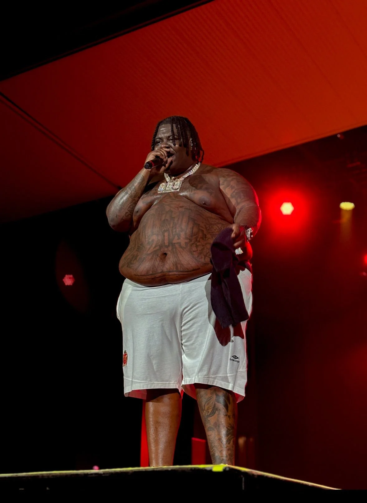 A shirtless man with tattoos standing on stage, holding a microphone, with a red lighting background.