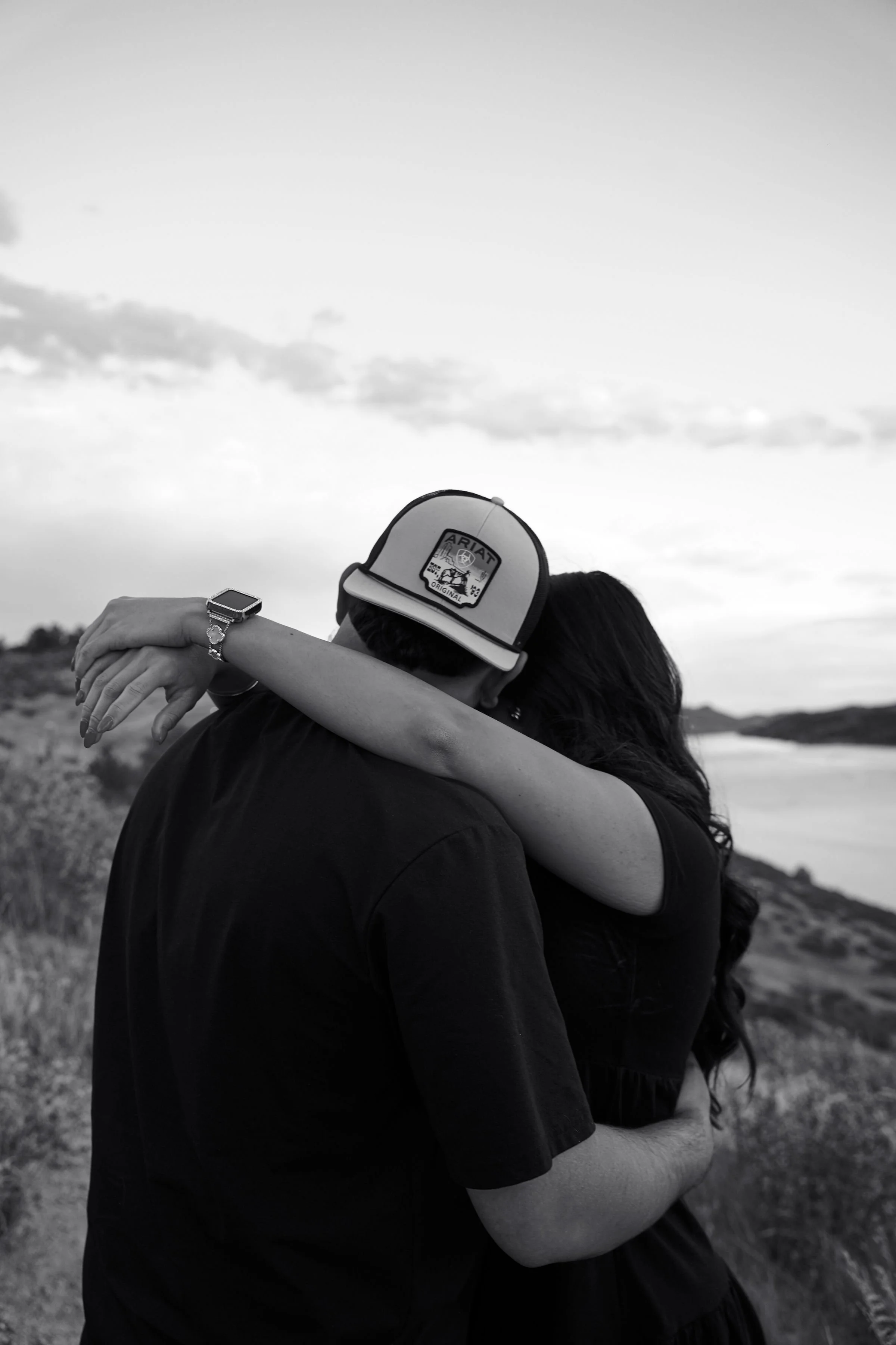 A black and white photo of a couple embracing outdoors, with the woman's arms around the man's neck, near a body of water and rolling hills.