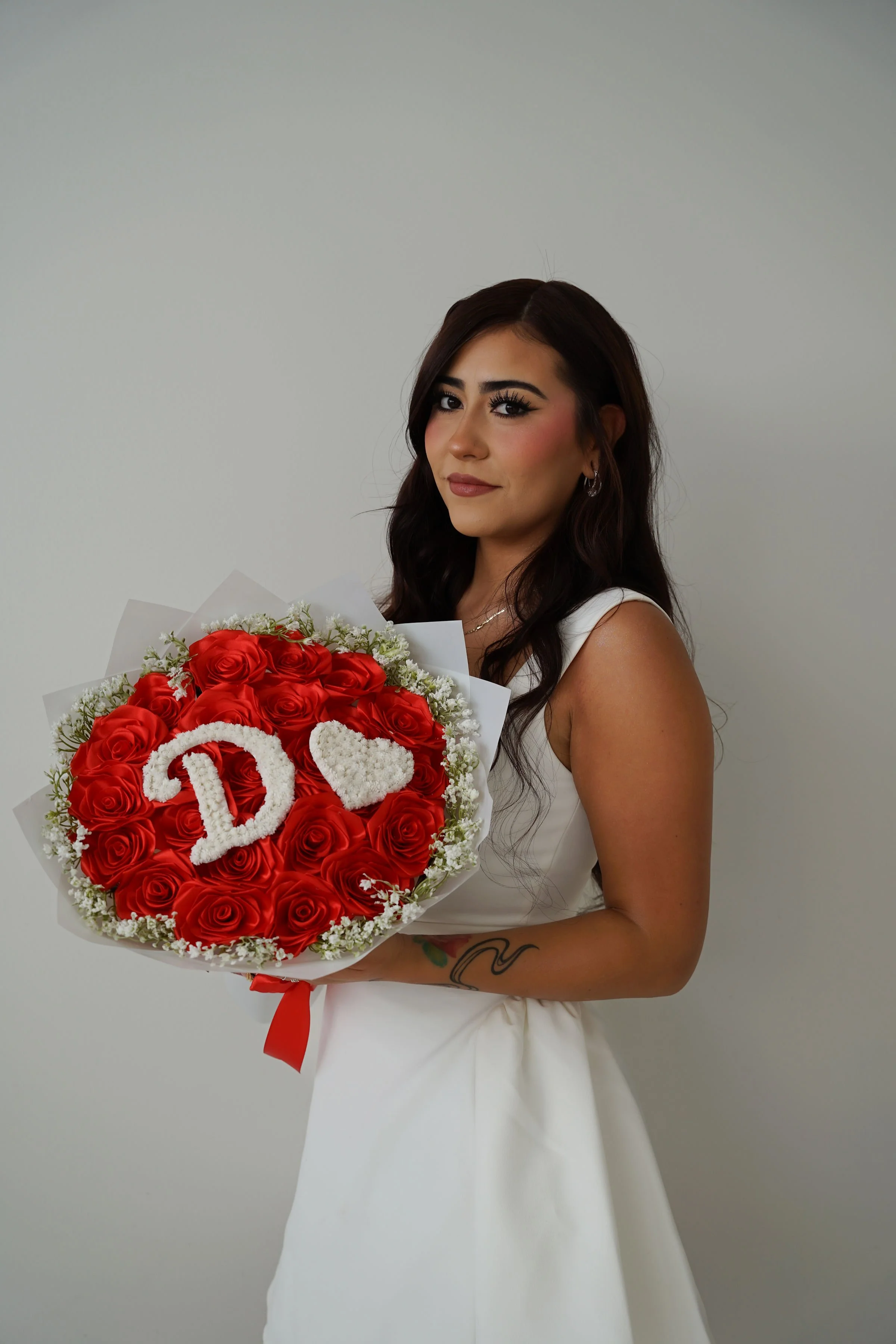 A woman in a white dress holding a bouquet of red roses with a white 'D' and a heart symbol made of flowers.